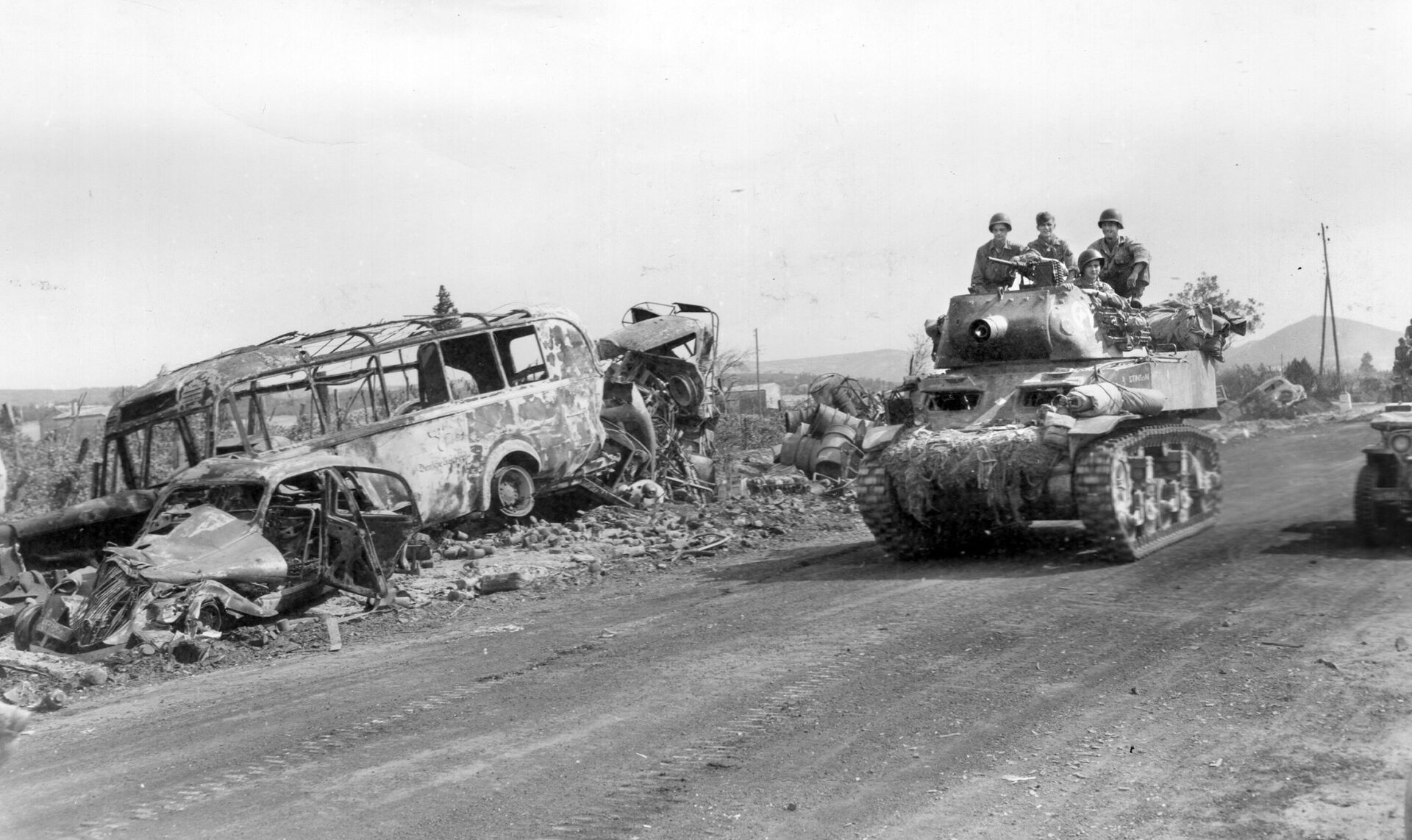 An American M8 gun carriage—an M5 Stuart light tank modified to carry a 75mm howitzer—rolls past the charred wreckage of a bus caught in the fighting along a road near Montelimar.