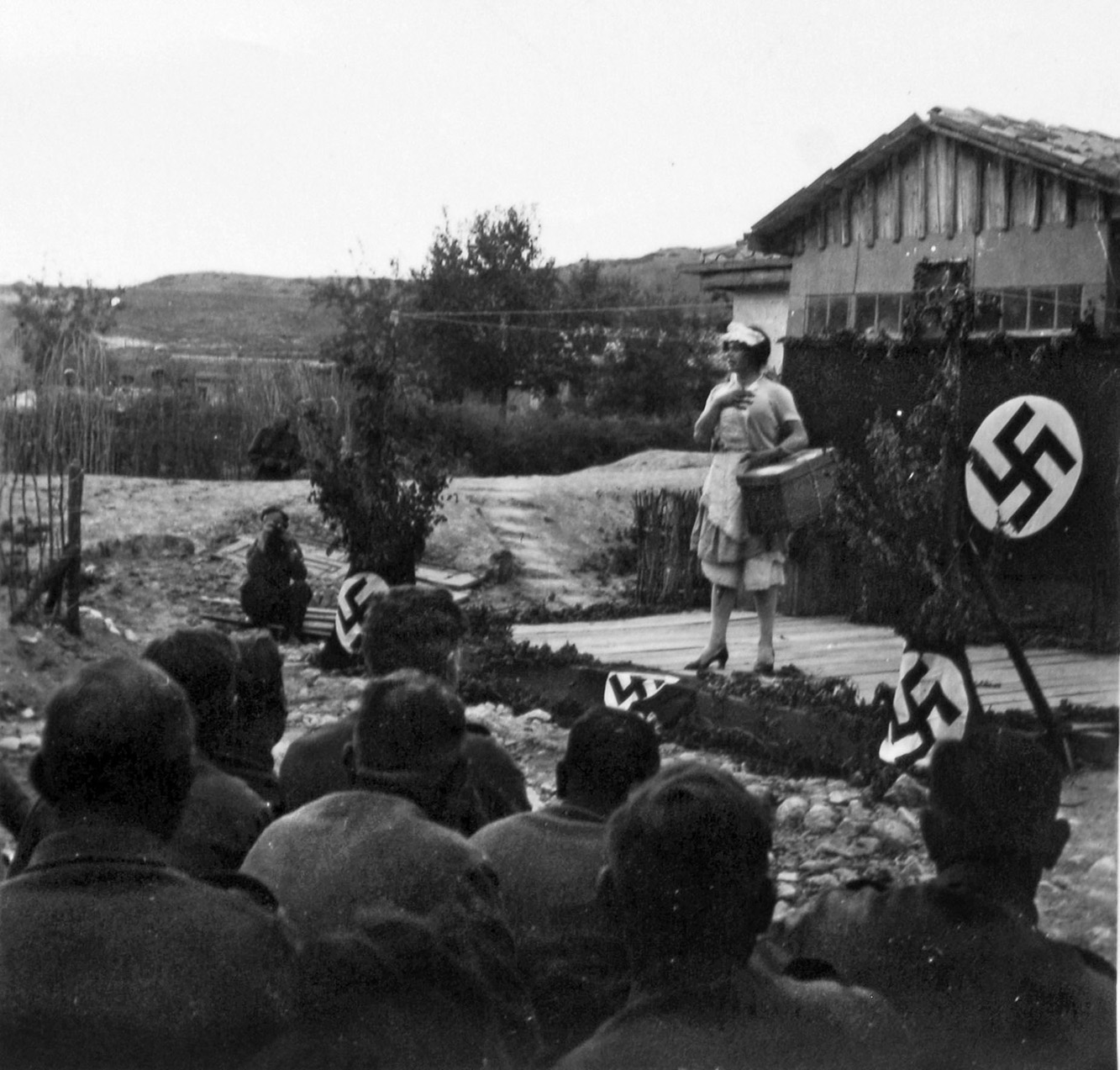 Appearing on the Eastern Front, a member of a traveling troupe of entertainers (the German version of the USO) performs on a makeshift, swastika-decorated stage. The performer is a male, not quite a female impersonator, but rather a staple of the humorous fare to which the troops were accustomed.