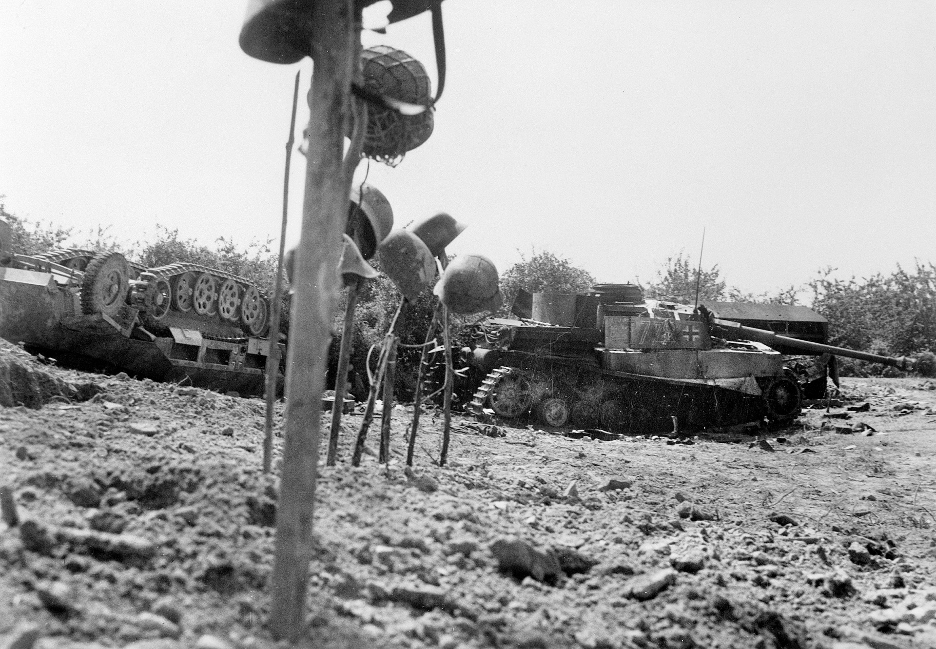 The graves of a German tank crew, killed in action in the Falaise Pocket, are marked by their helmets. In the background an overturned halftrack and a destroyed PzKpfw. IV medium tank bear mute testimony to the ferocity of the fighting.