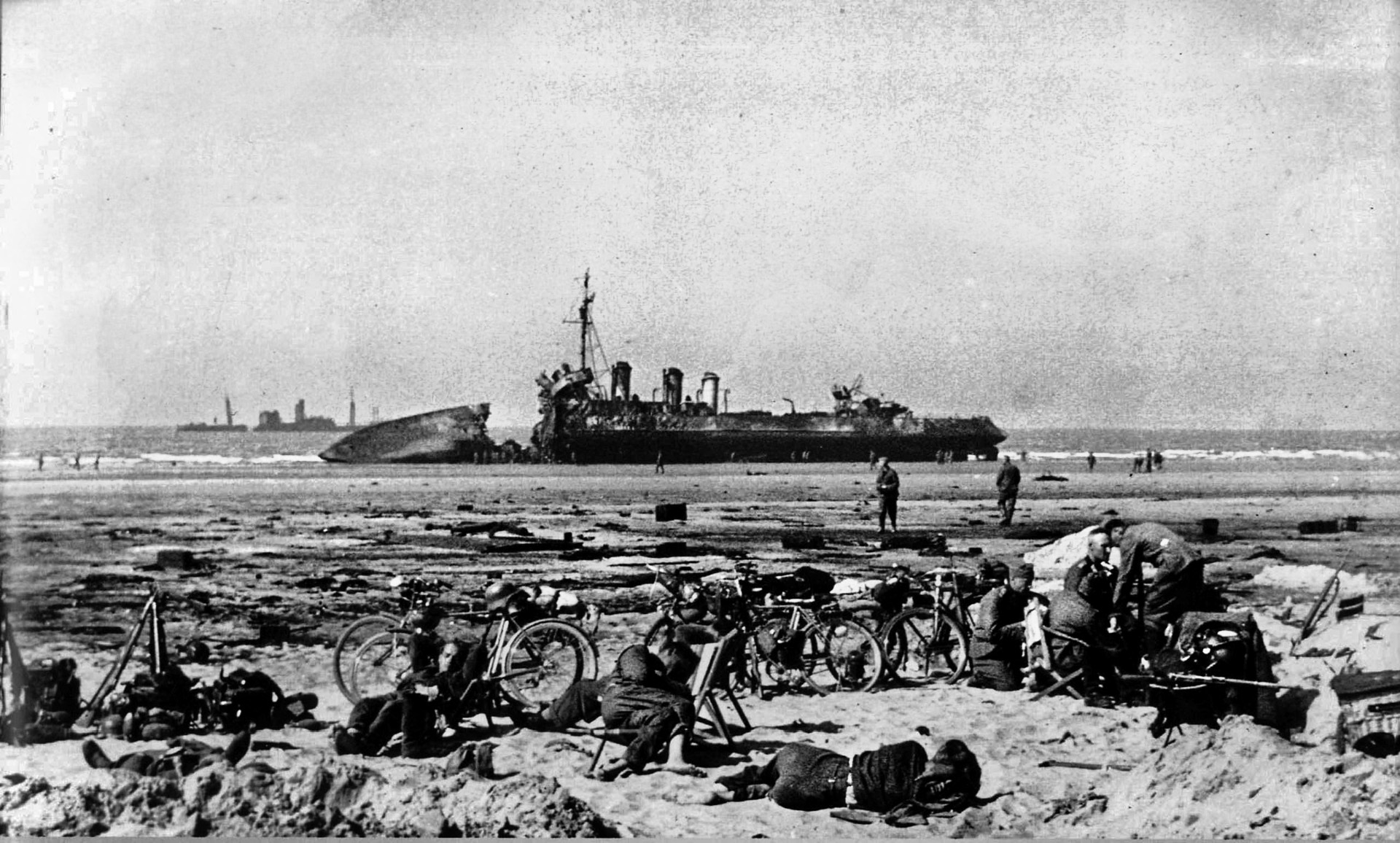 The French destroyer L’Adroit lies a twisted wreck off the shore of Dunkirk while German soldiers relax on the beach in the foreground following the withdrawal of the British Expeditionary Force.