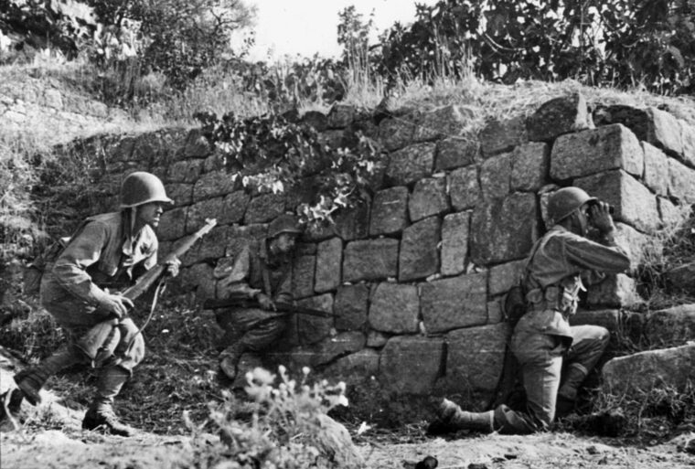 U.S. infantrymen take cover behind a wall in close-quarters fighting with Axis troops. The rough terrain of Sicily furnished fanatical German infantry with superb defensive positions.