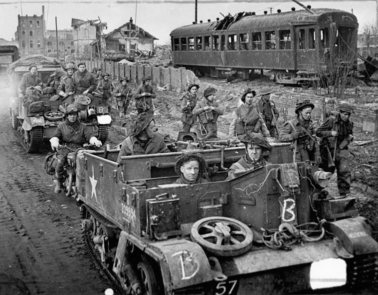 Gutted streetcars lie derelict along a rail siding as British soldiers, some walking and others aboard Bren Gun carriers, advance through the shattered streets of Goch on February 25, 1945, following the town’s capture during Operation Veritable.