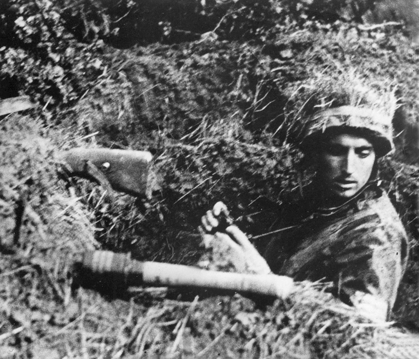 Although his cause is lost, a camouflaged German paratrooper (Fallschirmjäger), equipped with rifle, stick grenade, and binoculars, waits for the Americans to arrive.