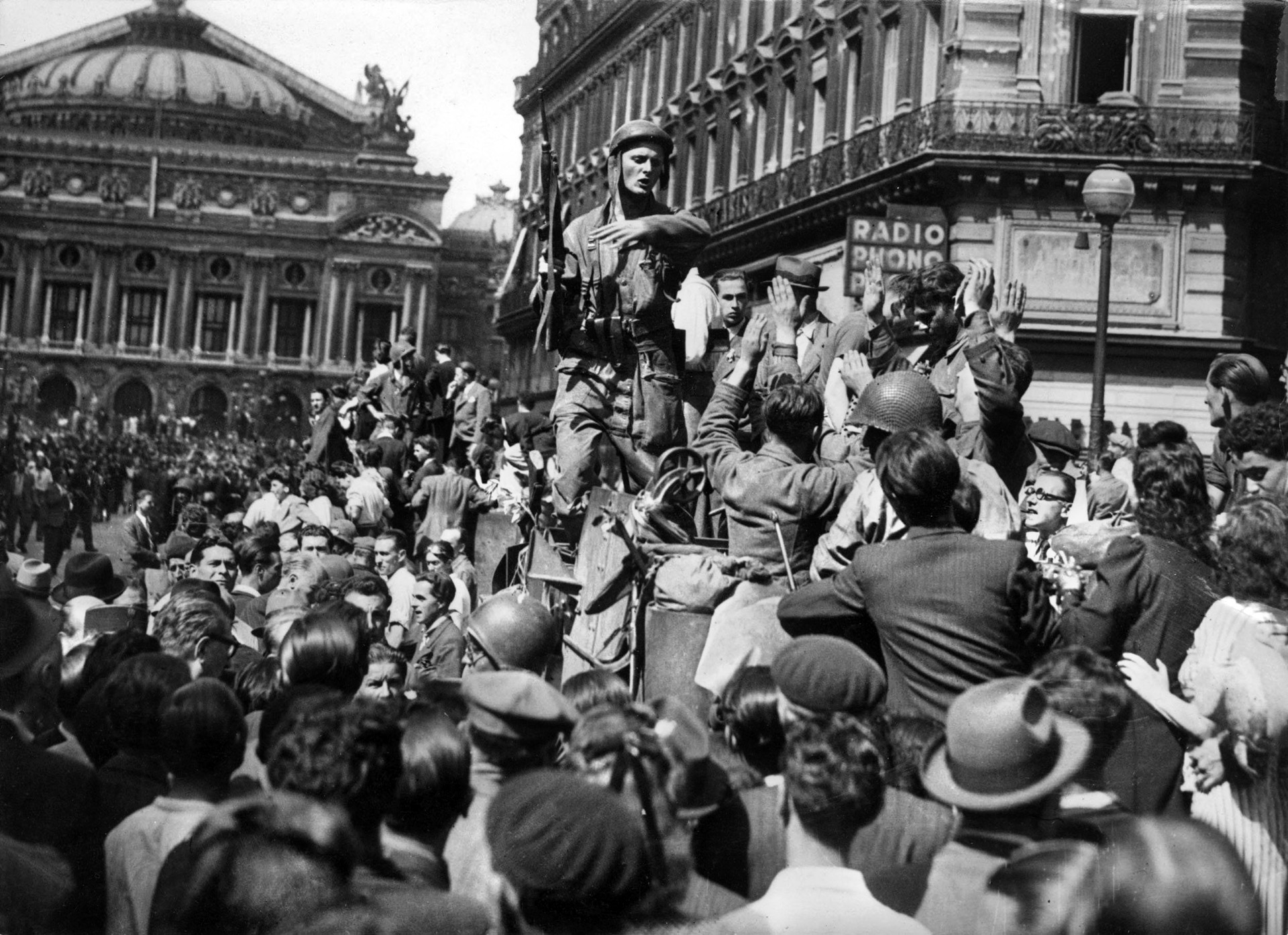 A French soldier with a carbine in his hand gestures in an attempt to keep Parisians away from German prisoners who are on his tank with their hands up. The Paris Opera House is in the background.