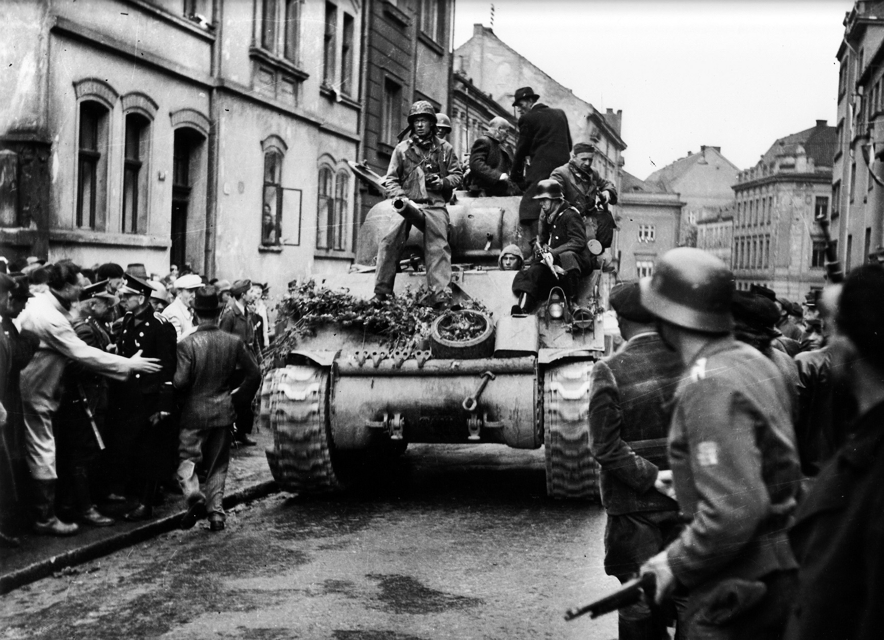 Local partisans in captured German uniforms watch as an American Sherman tank, with townsfolk on board, rolls through Pilsen following its liberation on May 6, 1945. 