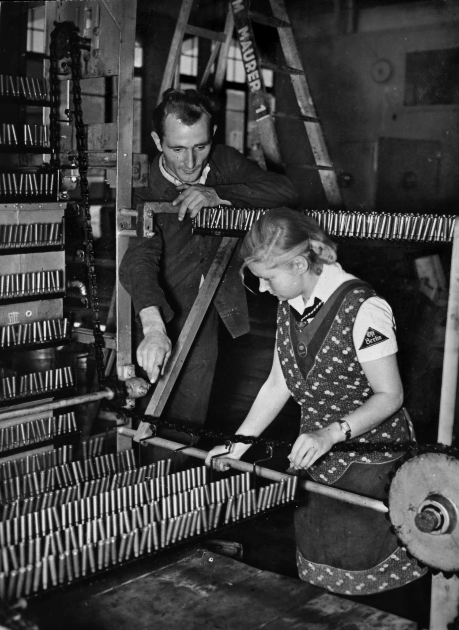 A BDM leader shows a young girl how to operate a piece of machinery on the floor of a German armaments factory in August 1942. Due to labor shortages, children were often pressed into service doing jobs that adults might otherwise perform.