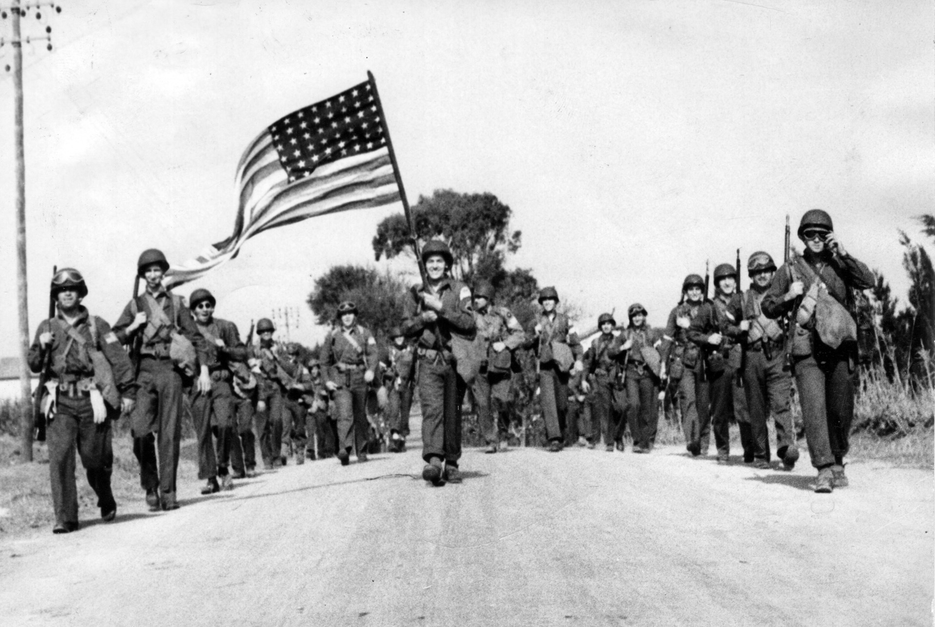 The 48-star U.S. flag catching the breeze at the head of their formation, U.S. paratroopers march toward the Maison Blanche airdrome, an installation near the city of Algiers. Maison Blanche served as a base for further airborne operations following the initial American drop during Operation Torch.