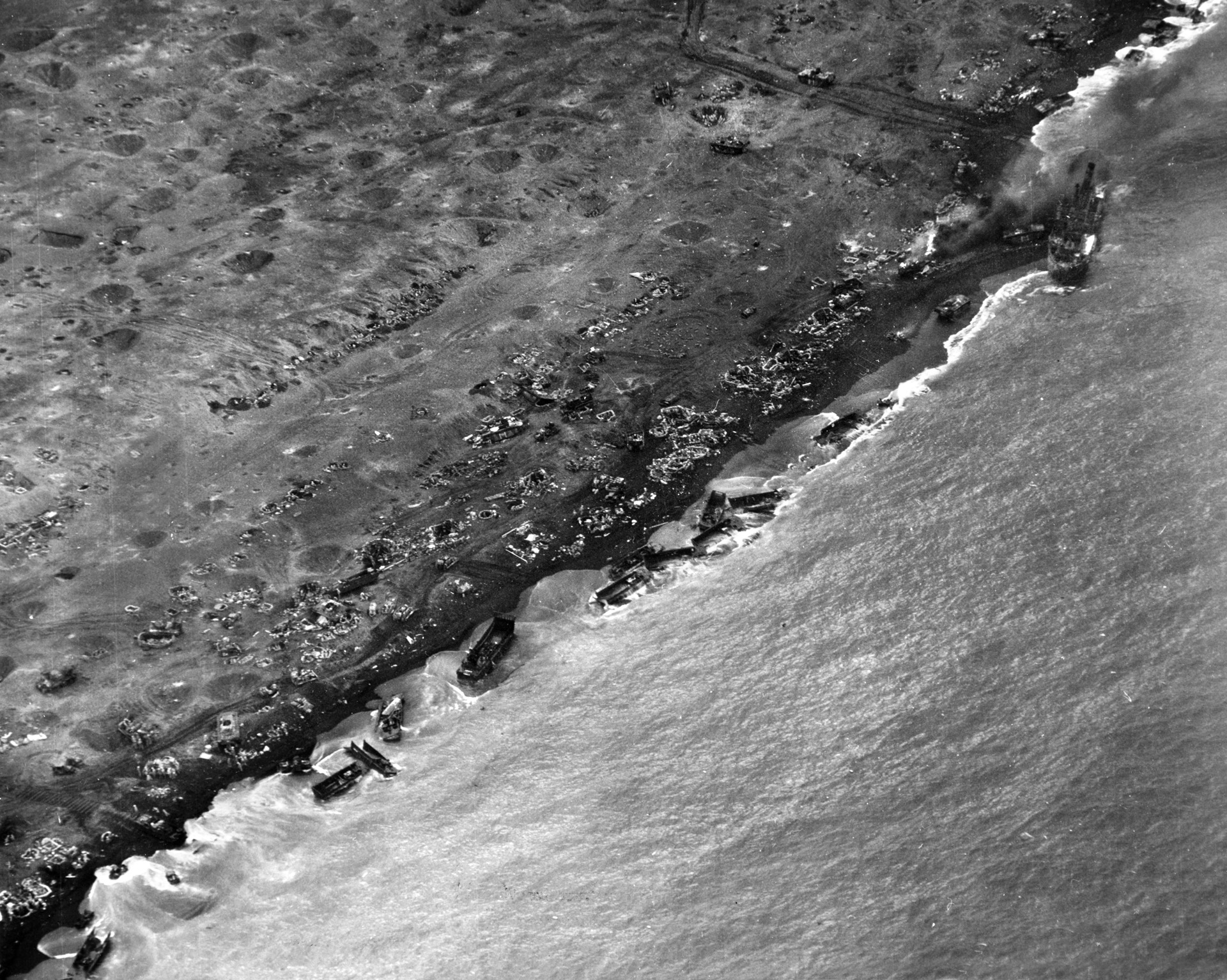 Wrecked landing craft litter the shore line at Iwo Jima, with shell craters and foxholes also visible in this aerial photograph. The Japanese held fire until the beach was crowded with American men and machines, while strong tides and surf also played havoc with the landings. 