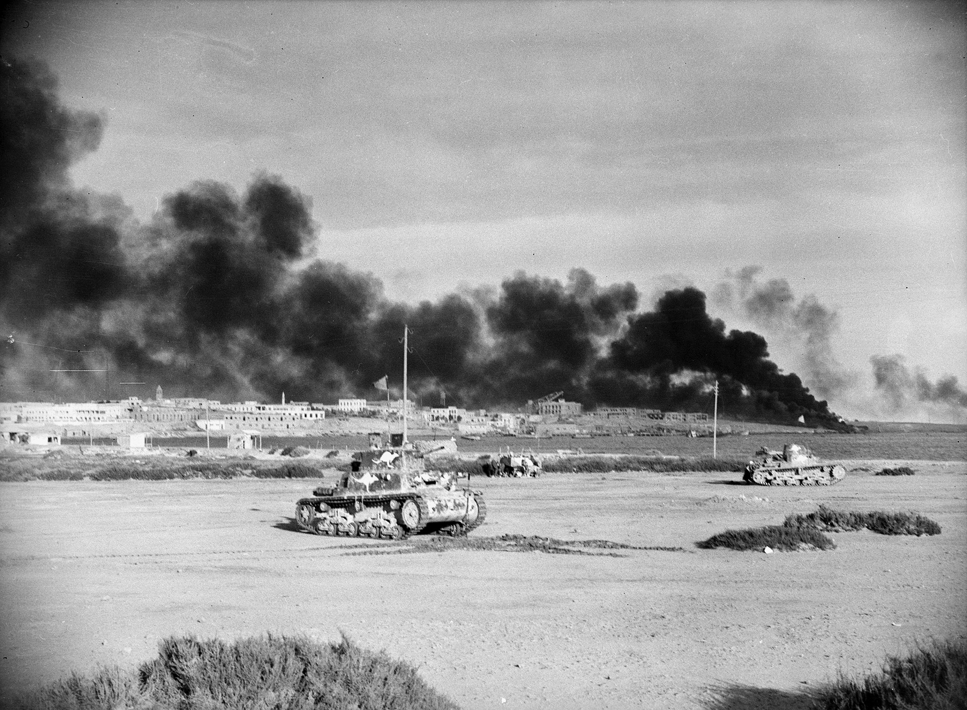 The port of Tobruk burns after its seizure in January 1941 by Allied forces. In the foreground are captured Italian tanks painted with white kangaroos to indicate they belong to the 6th Australian Division.