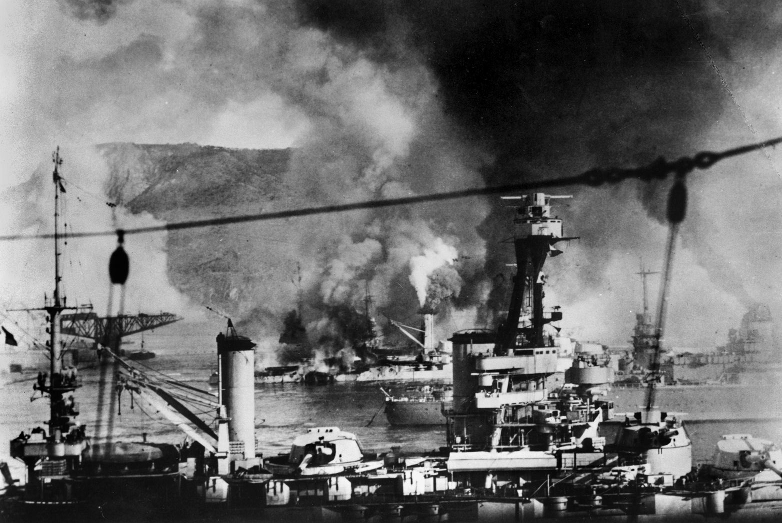 The elderly French battleship Provence is shown under fire in the foreground, while the modern  battleship Strasbourg escapes toward the open sea at right, and another old battleship, Bretagne, burns furiously in the background during the Royal Navy attack on the French fleet at Mers-el-Kebir.