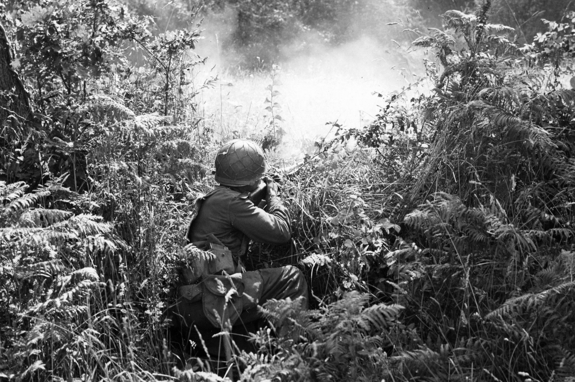 An American soldier, hidden in the Normandy terrain, fires on some Germans. At Brecourt Manor the Airborne troops utilized the German trenches for protection while they captured each artillery gun.