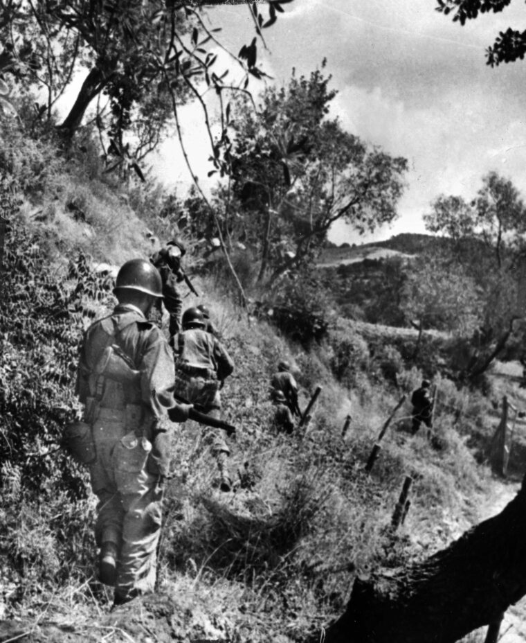 Crossing a steep embankment somewhere in Sicily, American infantrymen advance single file as they pick their way through a vineyard.