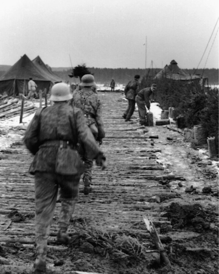 Two members of a Luftwaffe field division advance into an abandoned American camp as comrades secure jerry cans of fuel.