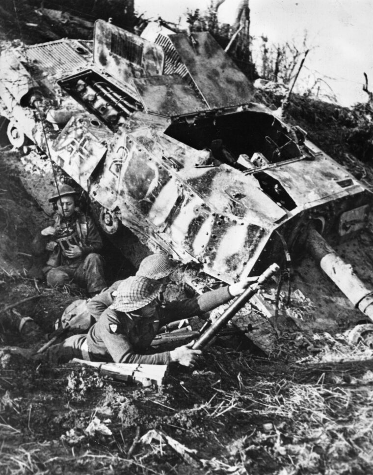 Polish soldiers, wearing British uniforms, fire a two-inch mortar while taking cover beside a destroyed German self-propelled gun. 