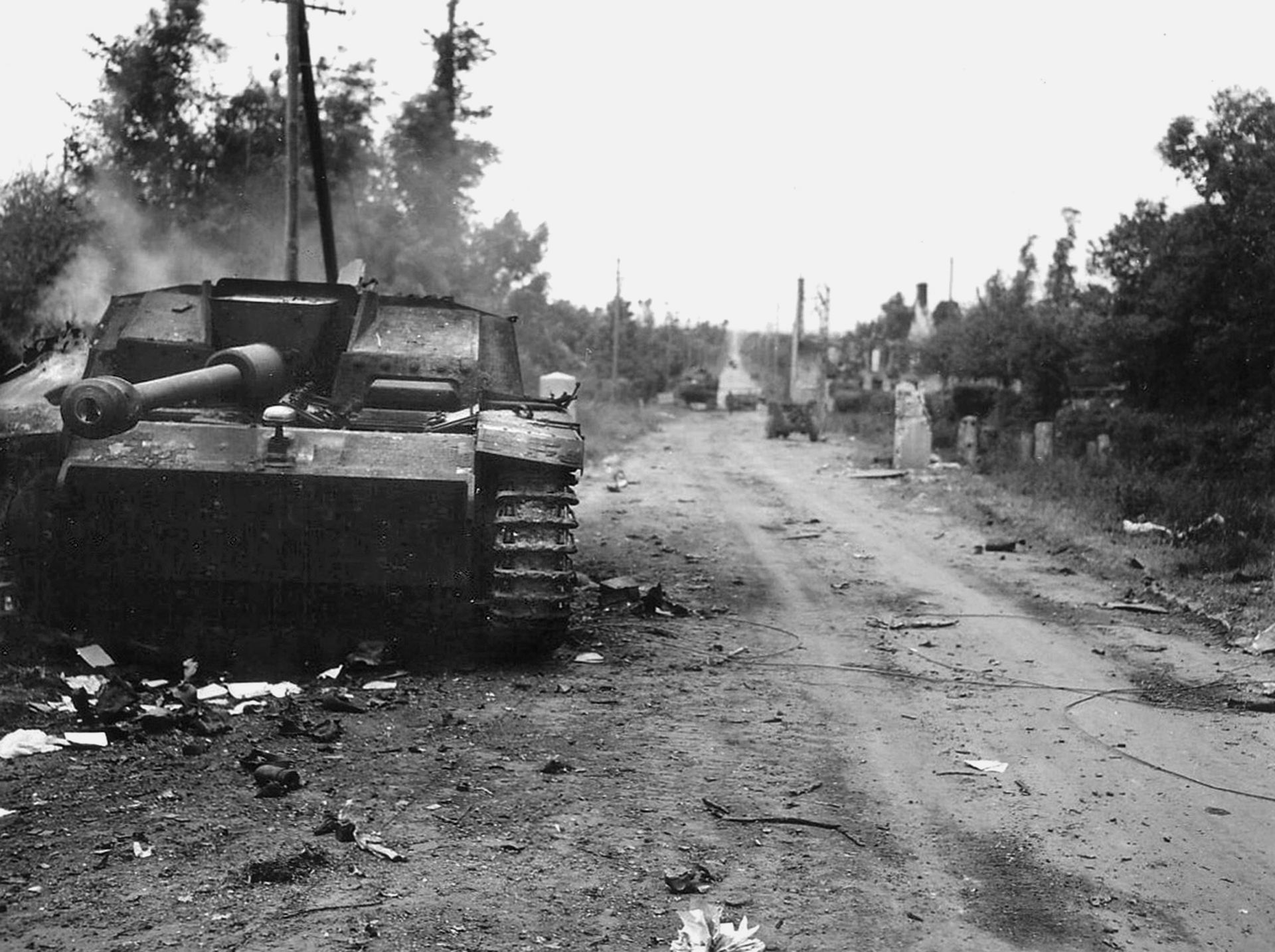 A destroyed German self-propelled gun smolders along the road leading from Neuville-au-Plain to Sainte-Mère-Église. Private John E. Atchley, H Company, 505th PIR, was credited with destroying the assault gun with a 57mm antitank gun—the first time he had ever fired one.  His courageous stand caused the Germans to halt their armored counter-attack against Sainte-Mère-Église.