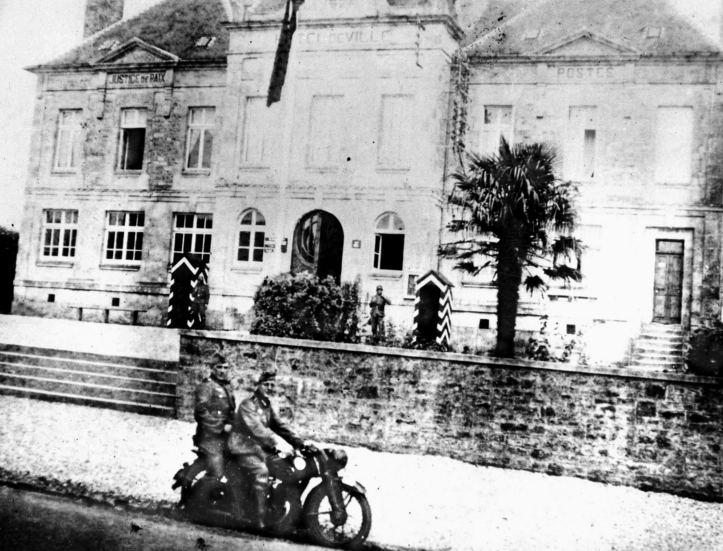 During the German occupation of Sainte-Mere-Eglise, two German soldiers on a motorcycle pose for the cameraman in front of city hall. 