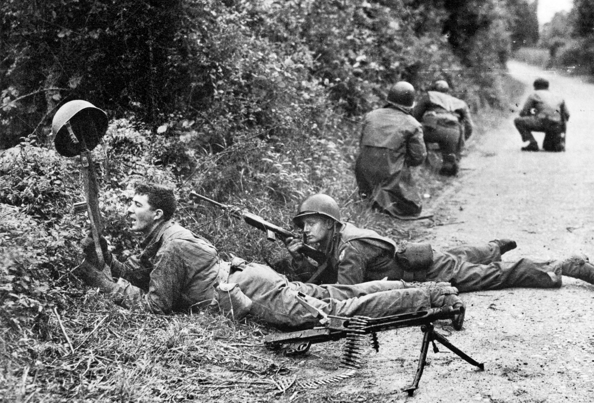 101st Airborne troopers guard a position along a farm lane in hedgerow country while a soldier holds his helmet up in the old trick of trying to draw fire so his buddy with a carbine can locate the enemy position.