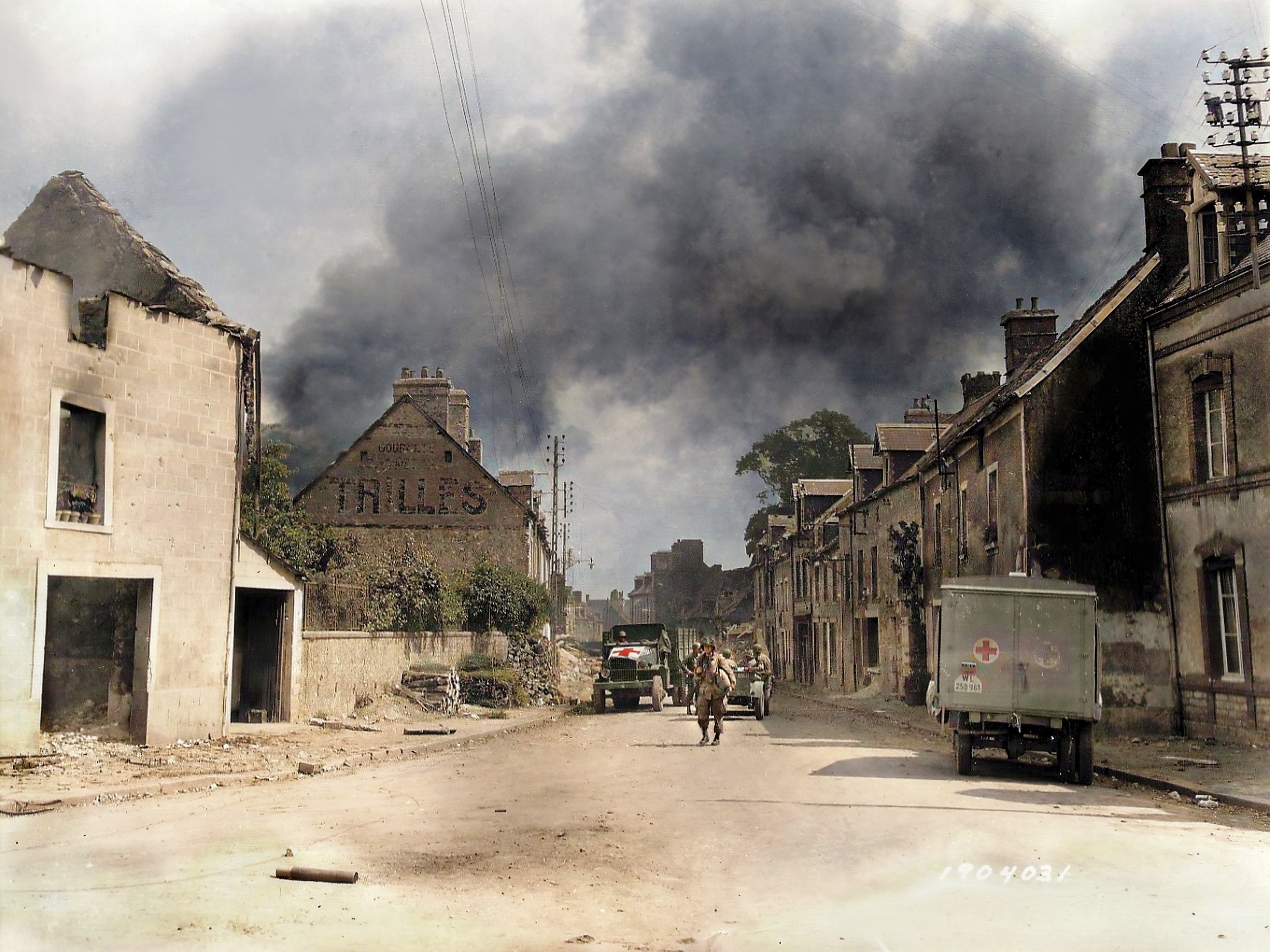 Smoke rises as paratroopers, ambulances, and a captured Kubelwagen populate a deserted street in Carentan in this colorized photo taken during the battle