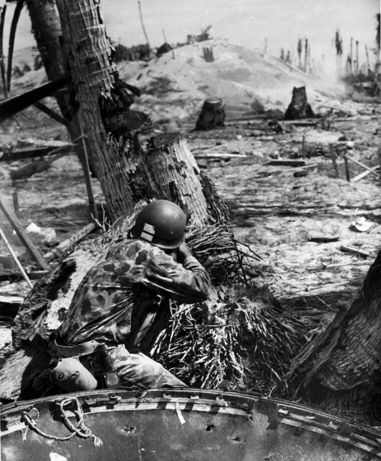 Crouched behind the remains of a tree, a Marine looks for a target behind a small opening on the Japanese bunker in the distance. 