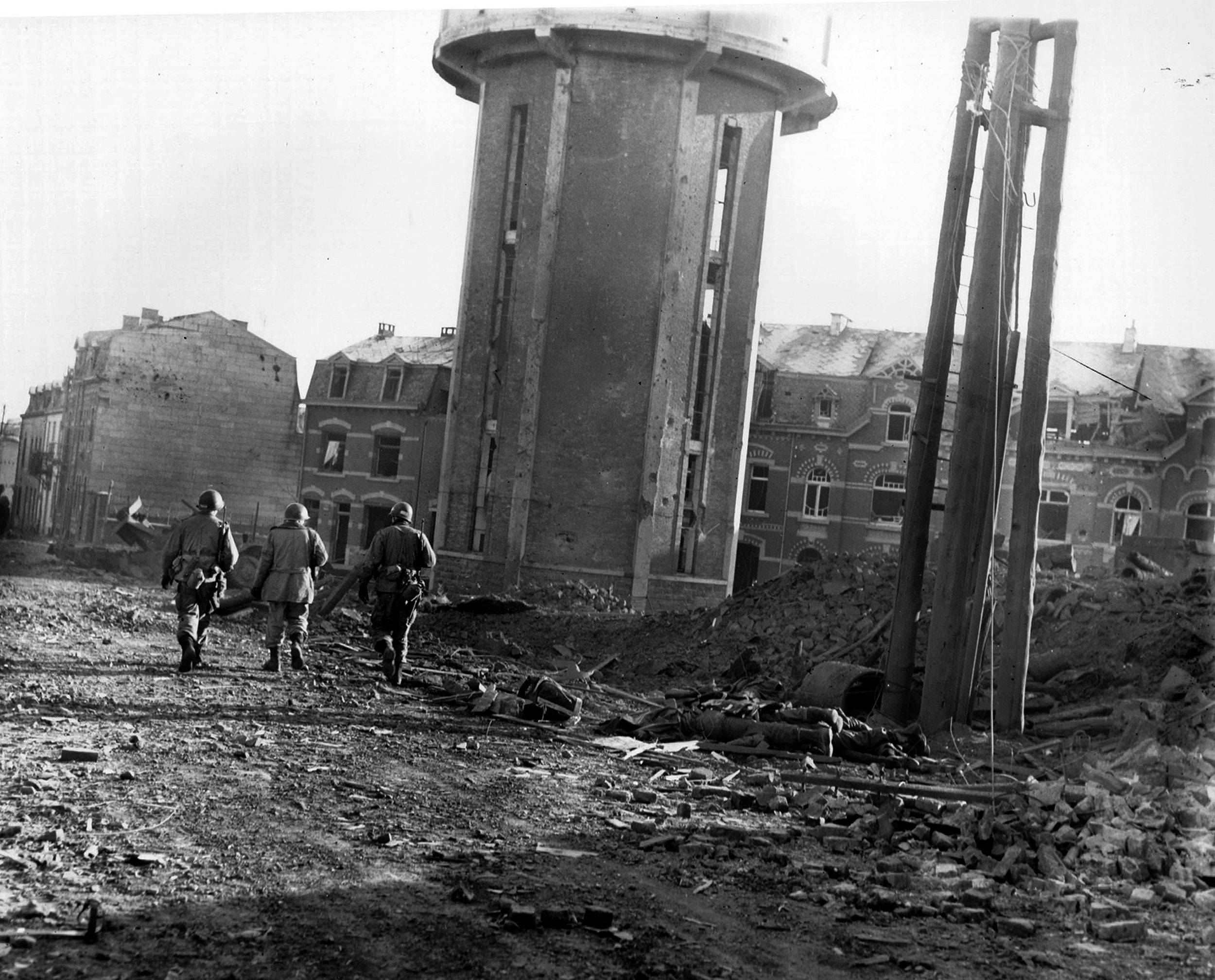 Their rifles slung over their shoulders, three men of the 101st Airborne Division walk down a rubble-strewn Bastogne street past the bodies of fellow soldiers killed by German bombing the previous night. This photograph was taken on Christmas Day, 1944, and the beleaguered defenders of the town were relieved the following day.