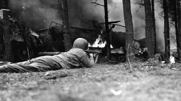In a wooded area near St. Vith an American soldier takes cover and trains his Thompson submachine gun on an enemy position. Savage fighting has already occurred in the area, as evidenced by the burning vehicle and debris littering the scene.
