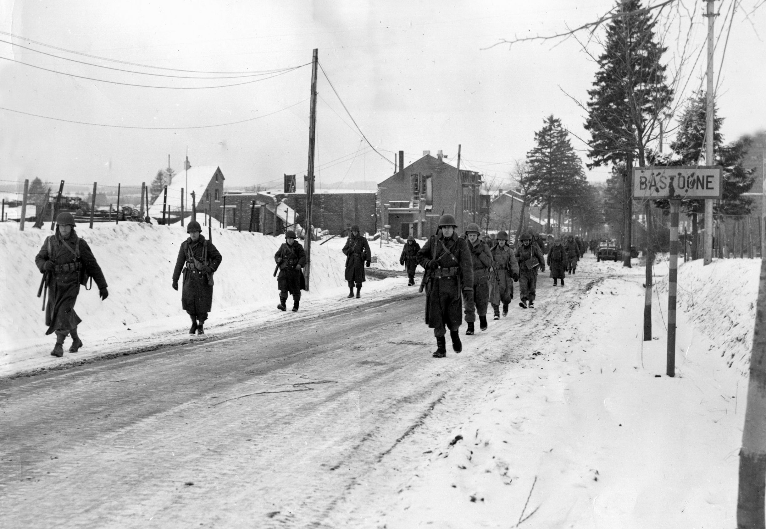 Members of a “Screaming Eagles” unit move out of snowy Bastogne on December 29, 1944, to attack enemy holed up in a nearby village. Note the bullet-scarred city limit sign.