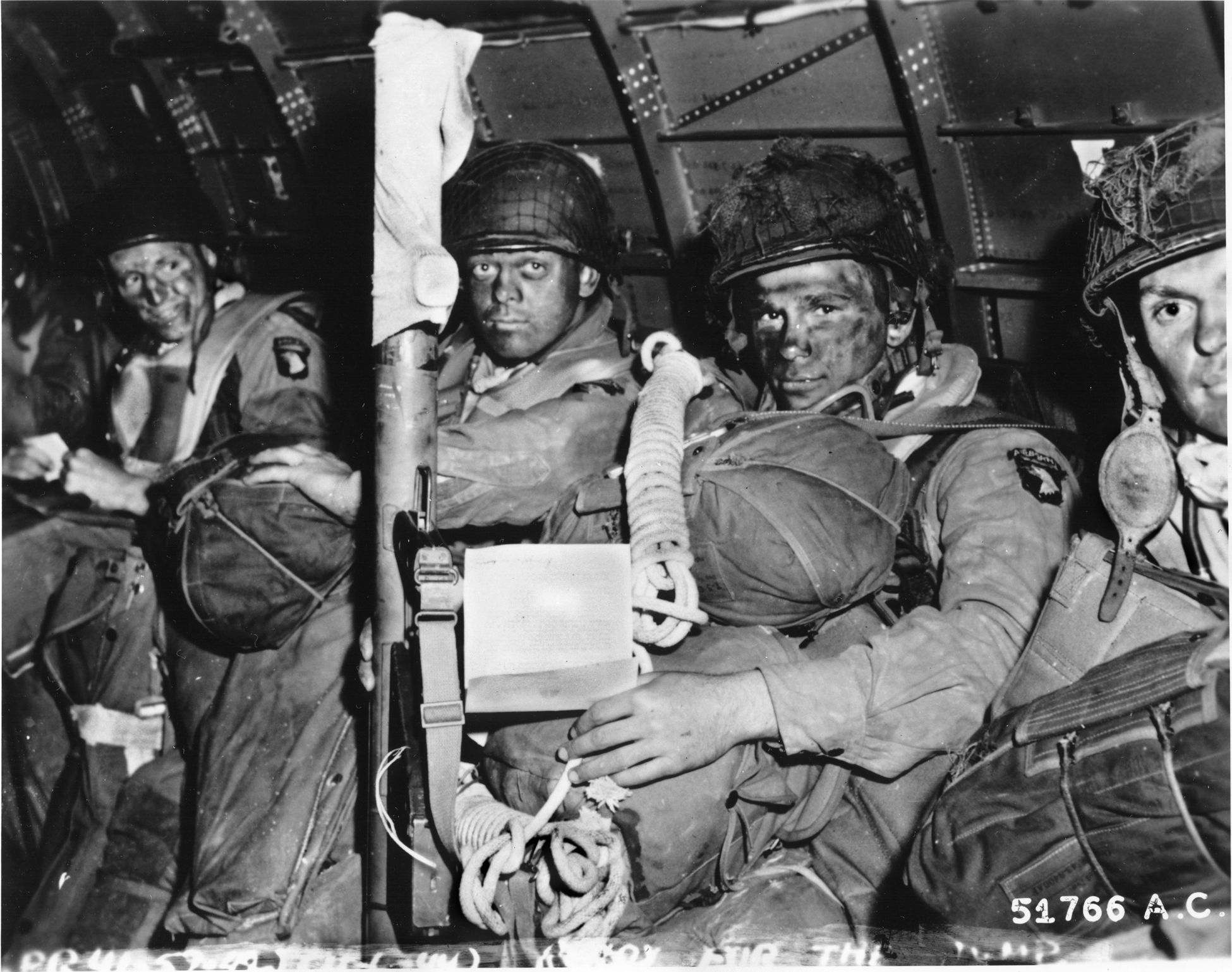 With blackened faces, a “stick” of 101st Airborne troops prepare to depart England. The bazookaman in front holds a copy of Eisenhower’s inspirational message for Operation Overlord.