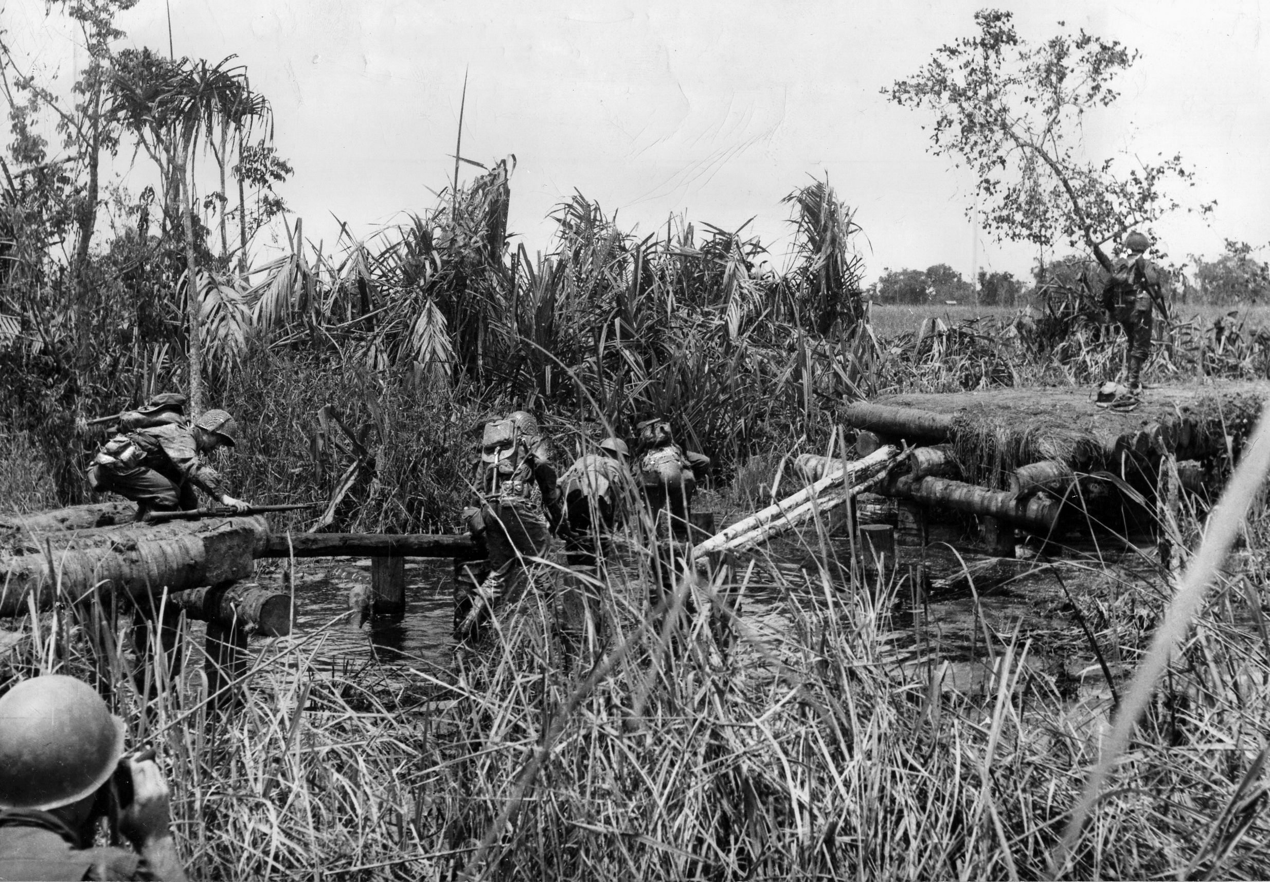Swamps, heat, humidity, poisonous snakes, disease-bearing insects, and impenetrable jungle added to the misery of the fighting. Here, American soldiers make their way across a bridge damaged by Japanese artillery and machine-gun fire during a phase of the capture of Simami Creek. 