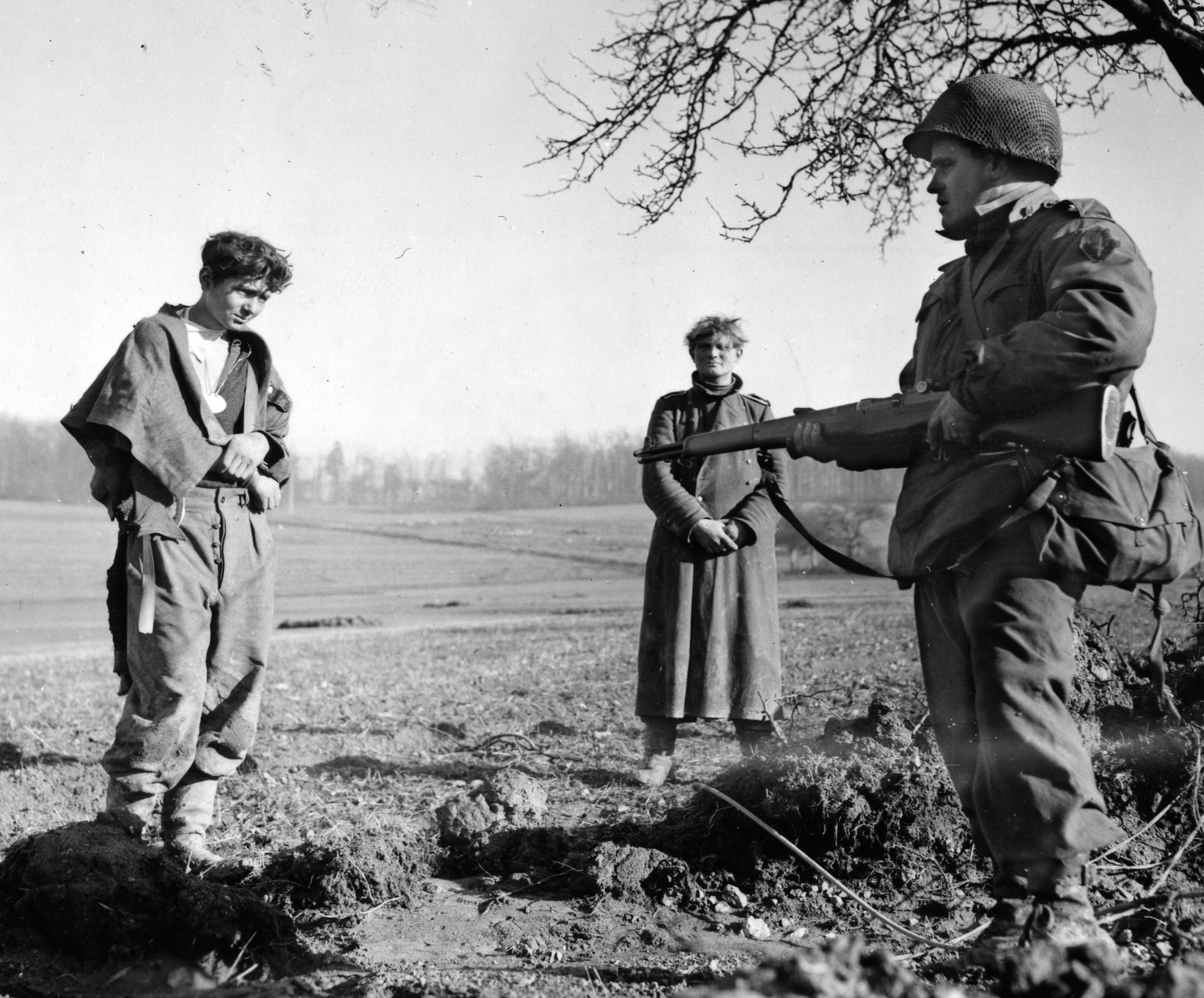 Two German prisoners stand under the watchful eye of an American soldier. These soldiers appear haggard and disconcerted after days of fighting and were captured while trying to infiltrate Allied lines. 