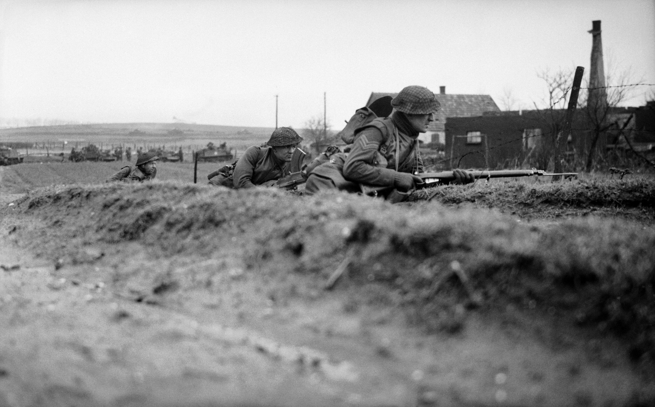 A British infantryman, his bayonet fixed, peers warily from a roadside as he prepares to move forward with comrades during the early stage of Operation Veritable.  Note the concentration of armored vehicles in the background.