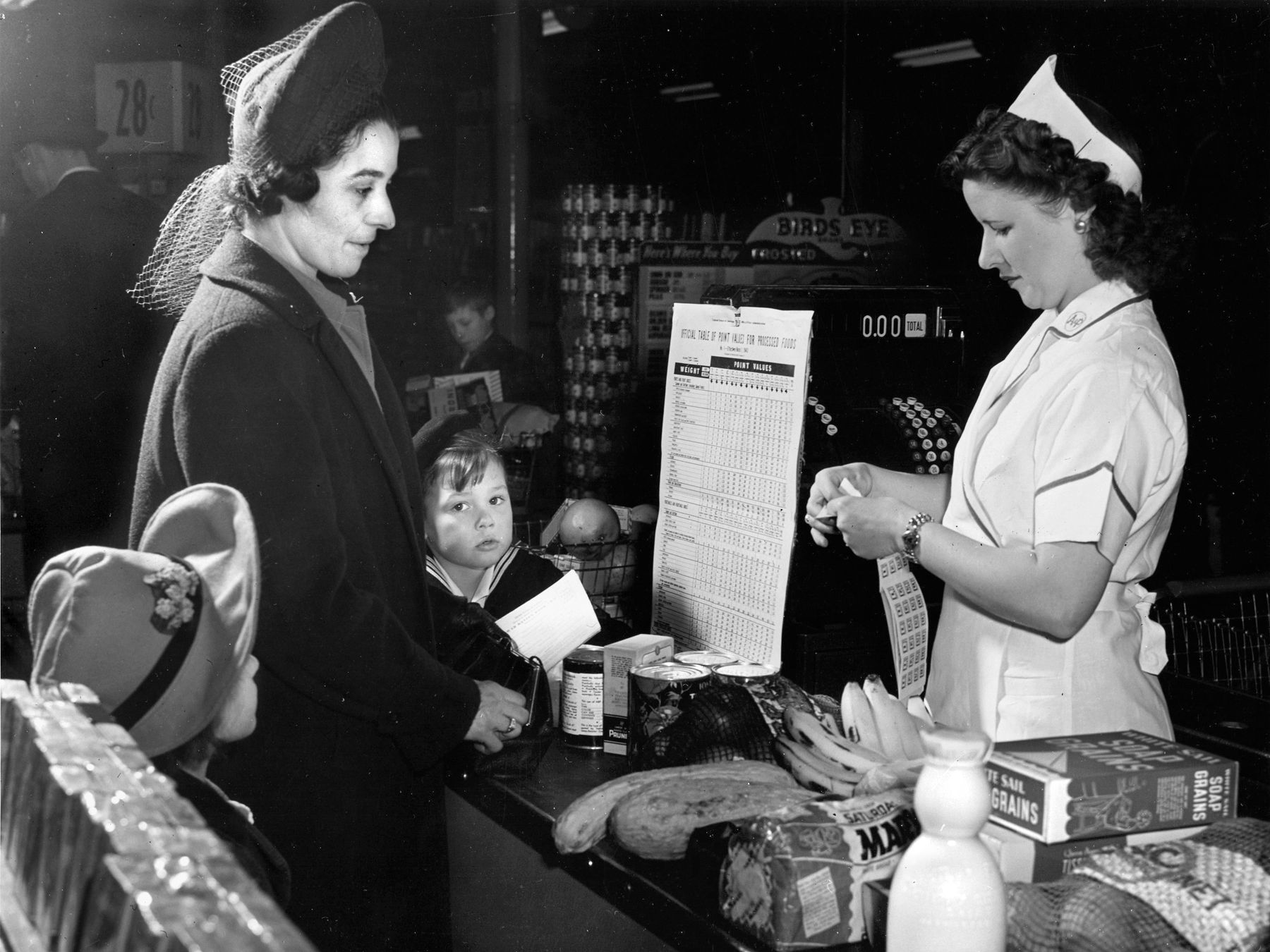 A shopper and her children watch as a clerk tears point stamps from a War Ration Book 2 to cover the processed foods being purchased. 