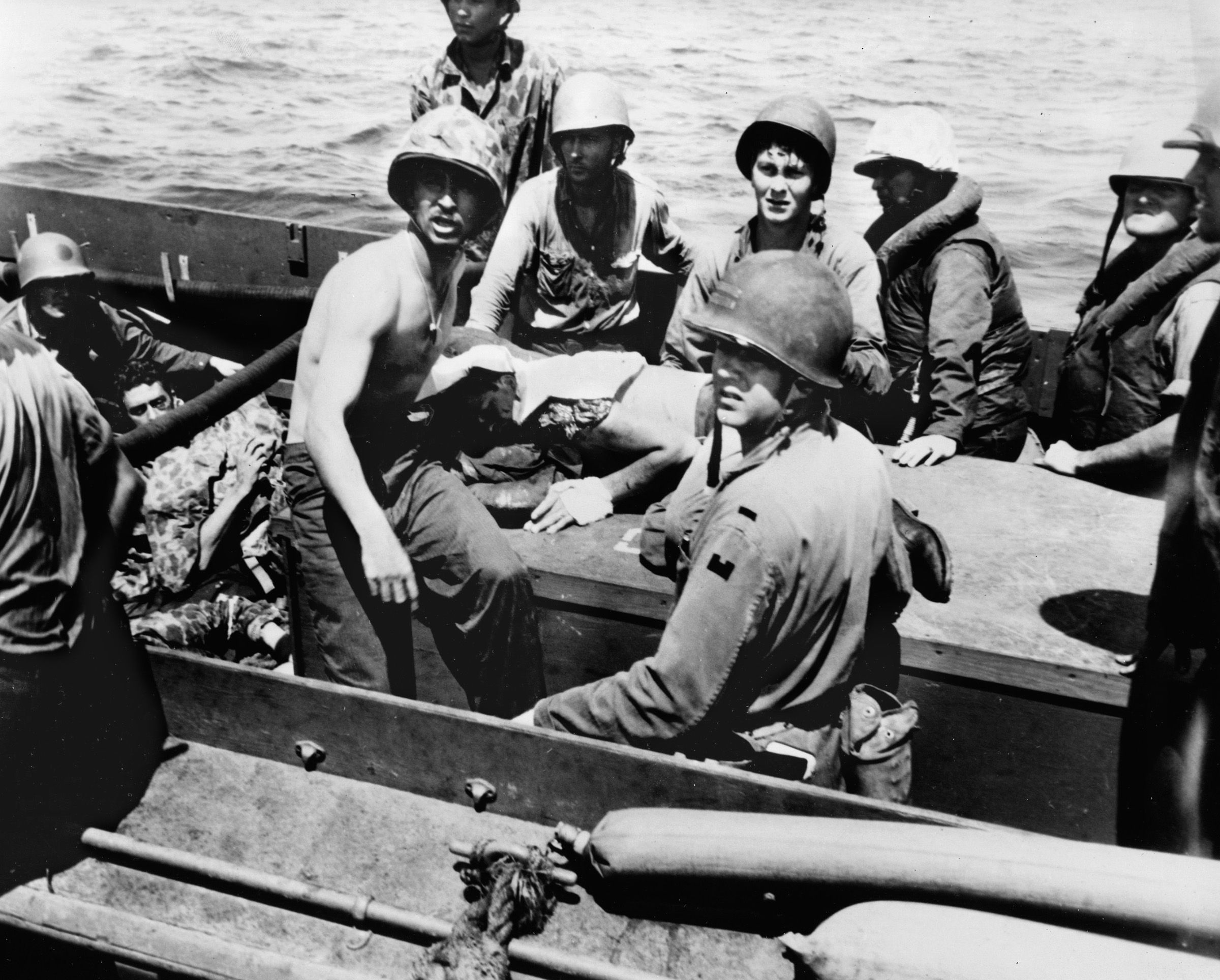 Wounded Marines are transported back to a troopship in a landing craft for transfer to a base hospital. One Marine lies on the bottom of the boat while another is on the hatch cover, his face covered to keep the flies off his wounds.