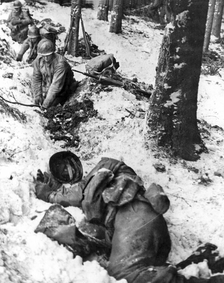American soldiers of the 110th Regiment, 28th infantry Division dig foxholes and remain in the line during the Battle of the Bulge. In the foreground, the body of a fellow GI killed in action lies in the snow.