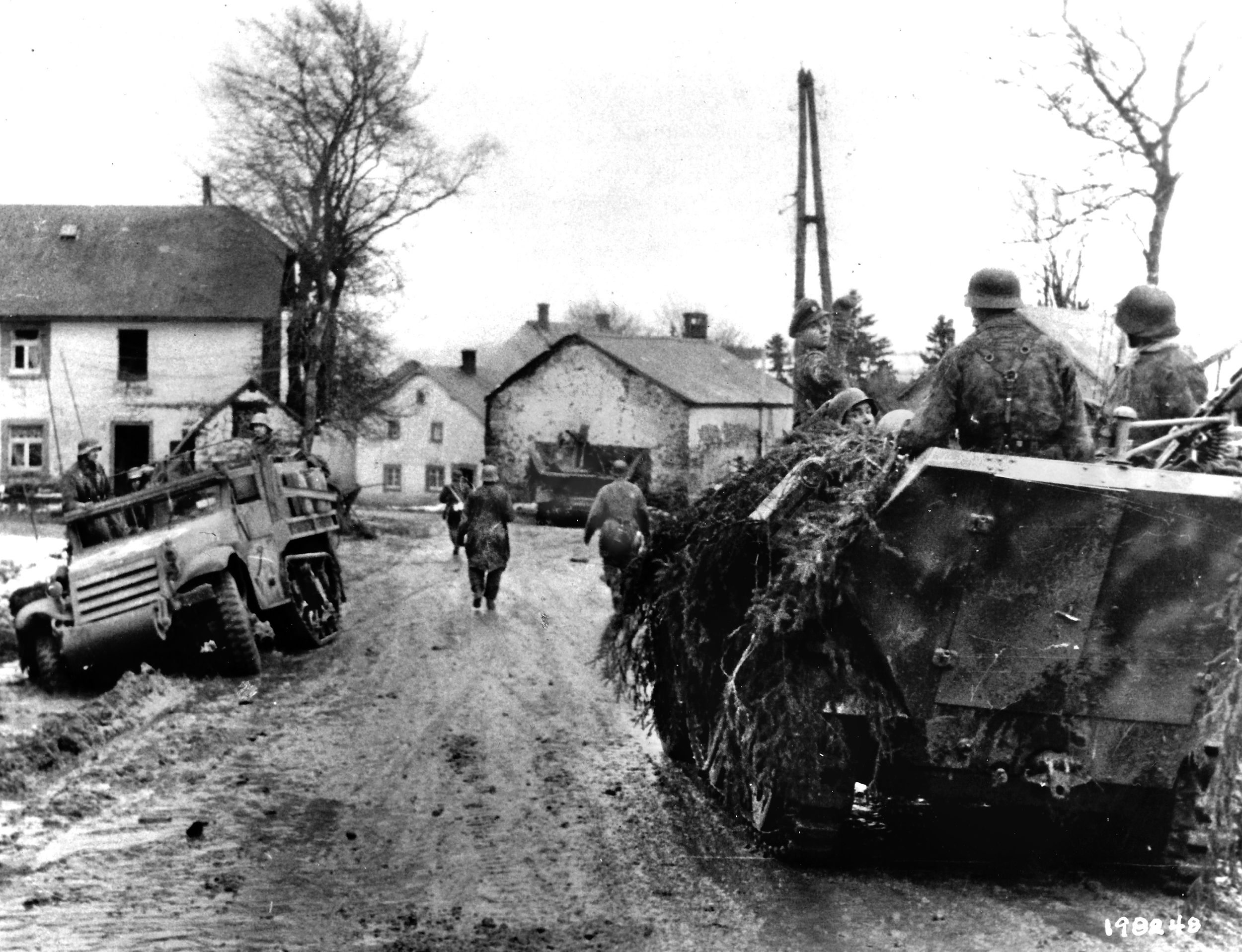 German infantrymen, on foot and riding in a halftrack, proceed through a village their forces have recently occupied. At left, German soldiers inspect an abandoned American M3 halftrack.