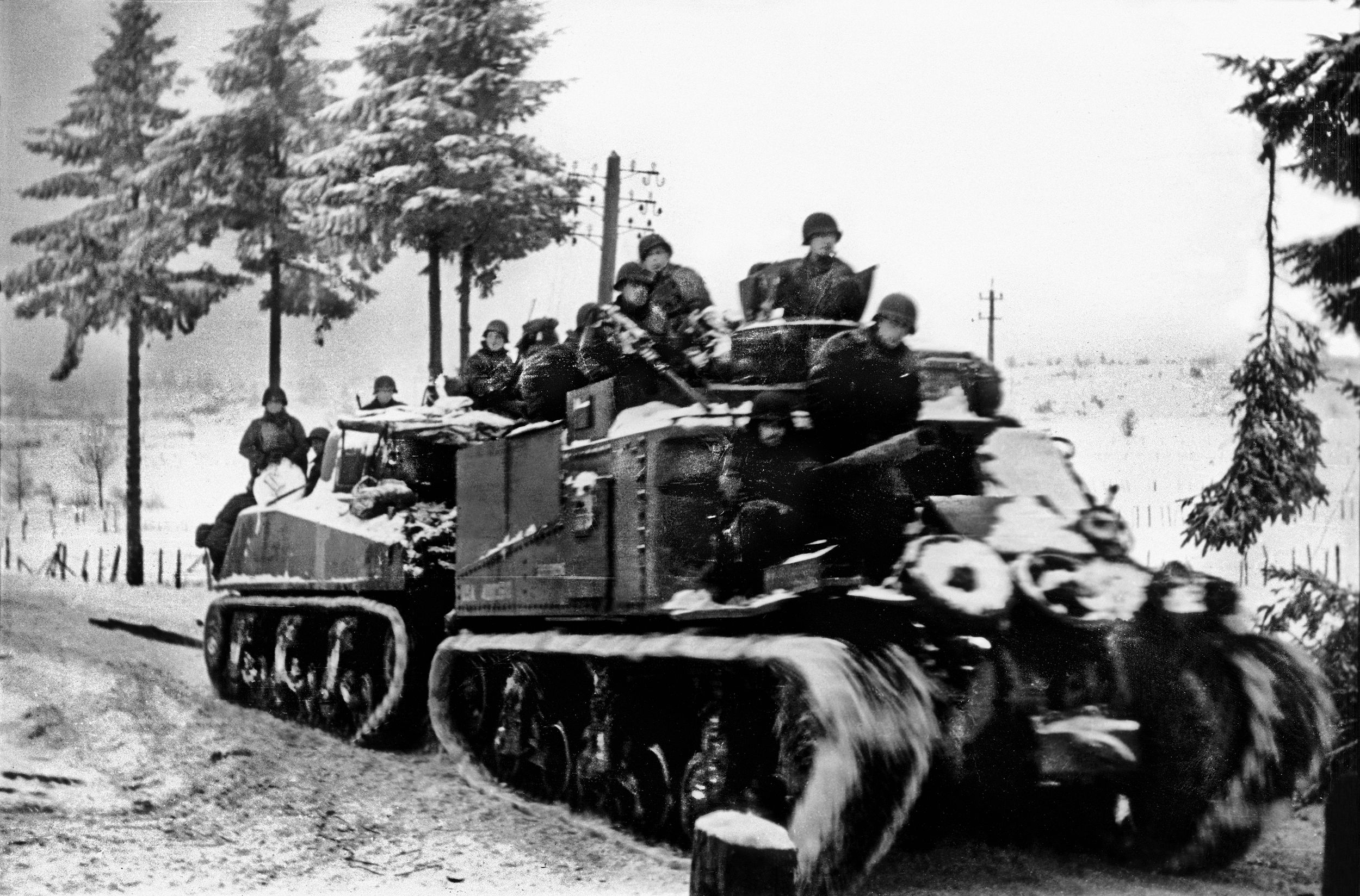 American soldiers hitch a ride on a pair of armored vehicles, one of them an M4 Sherman tank, during the Battle of the Bulge. On the afternoon of the first day of the German offensive, the American 707th Tank Battalion received orders to attack south along Skyline Drive and reached the village of Hosingen to bolster the defenders of Company K, 110th Infantry Regiment.