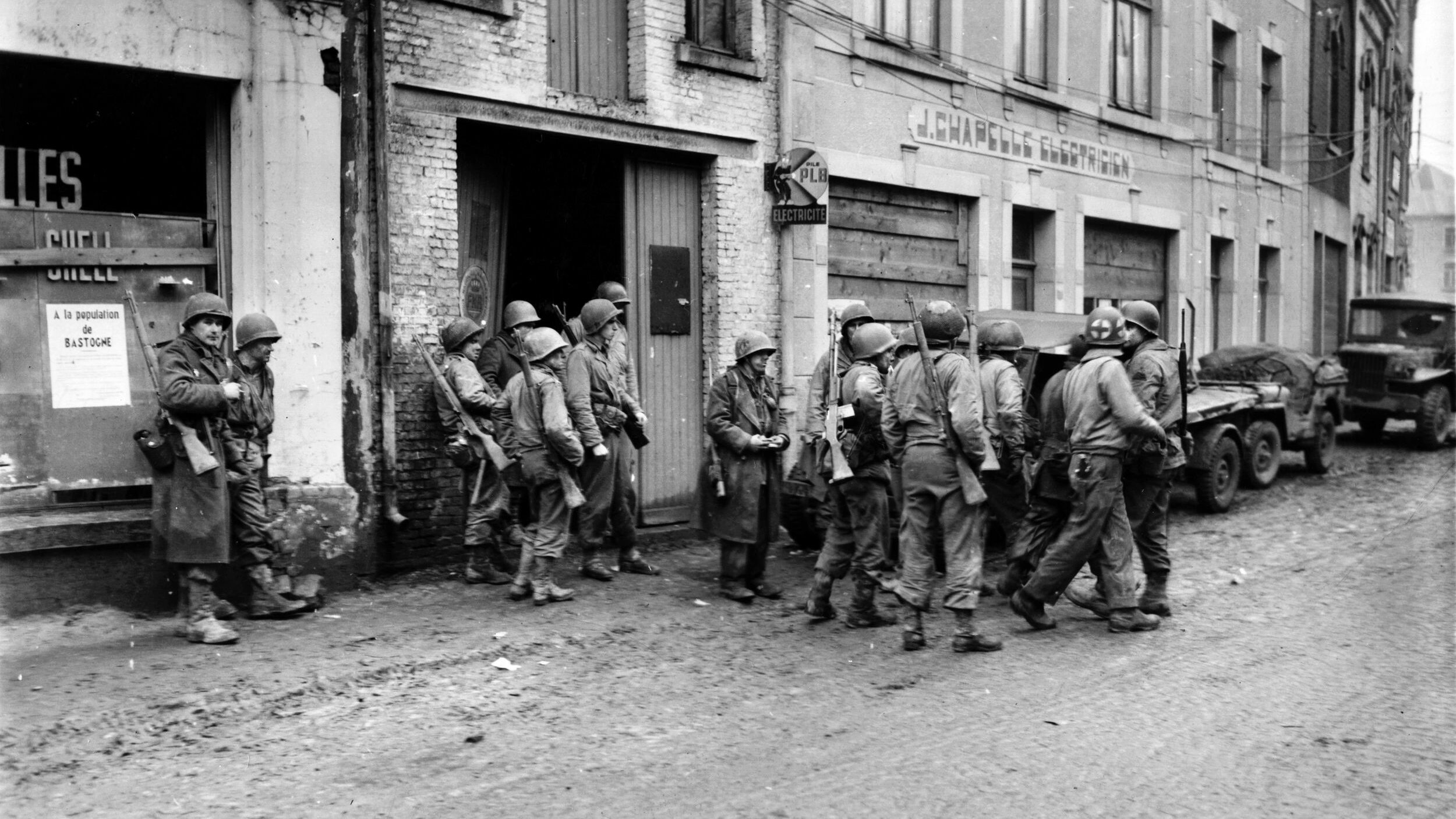 28th Division soldiers who avoided capture as their positions were overrun congregate in Bastogne. One of the soldiers is carrying a Browning Automatic Rifle (BAR), while others shoulder the M1 Garand, and M1 carbine.