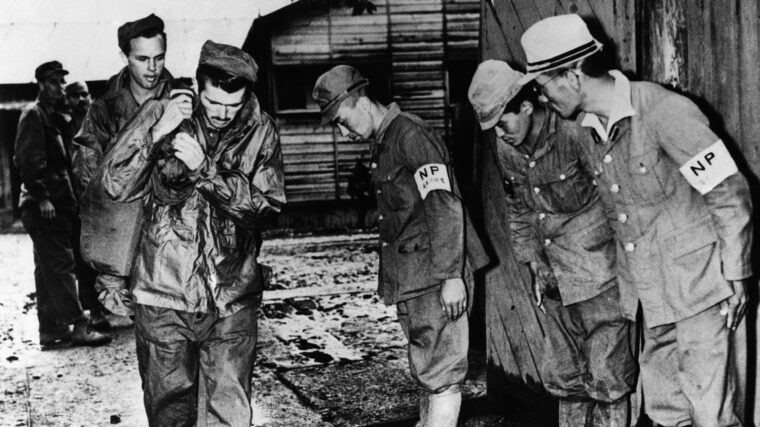 Exiting toward freedom, former Allied prisoners of war carry their belongings to waiting transportation as Japanese guards bow humbly. Thousands of Allied POWs were freed at the end of the war, but others met terrible fates aboard hell ships or were executed by their captors.
