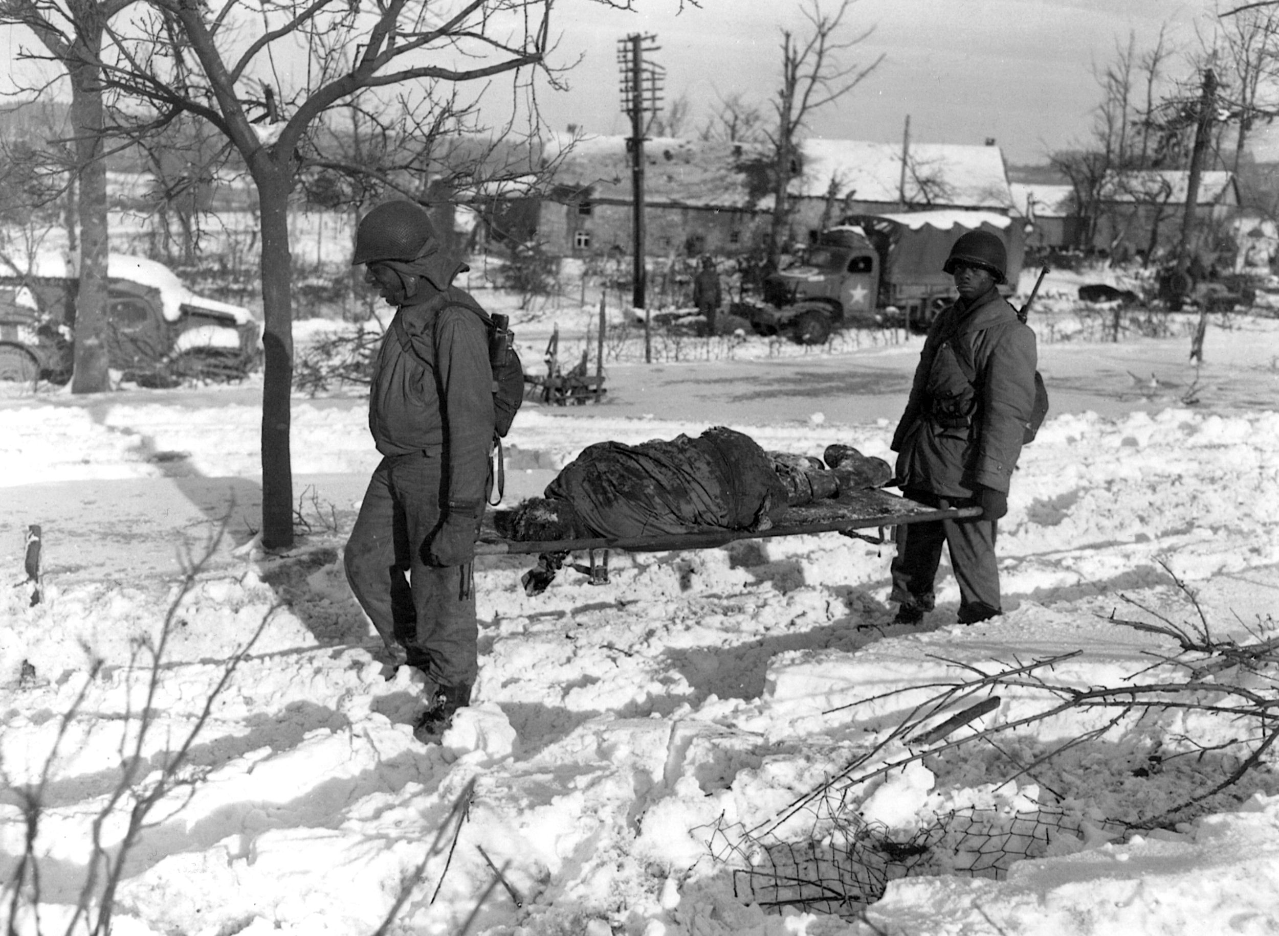 An American soldier, killed in the “Malmedy Massacre” during the Battle of the Bulge, is carried off the field by medical troops. 