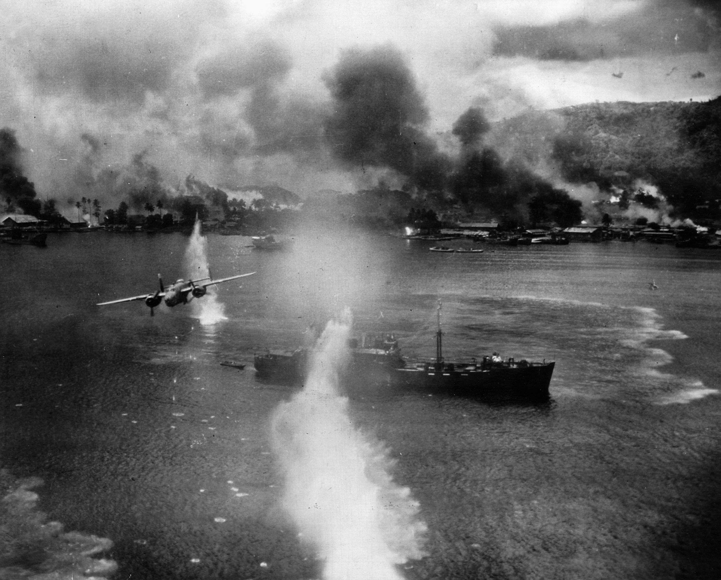 Black smoke hangs over shore installations as a U.S. B-25 bomber streaks above a Japanese merchant ship in the Rabaul harbor.