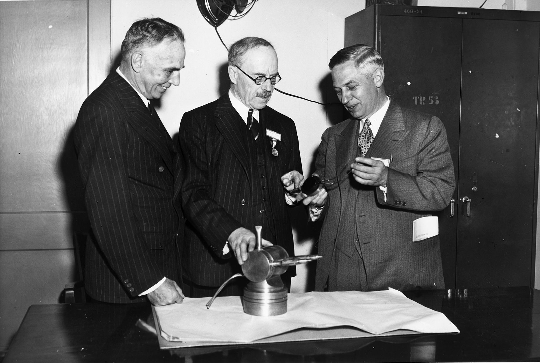 Henry Thomas Tizard, Alfred L. Loomis, and Lee Alvin-DuBridge examine a magnetron on a table in front of them in this image. The three scientists were involved in the development of airborne radar for the Allies during World War II.