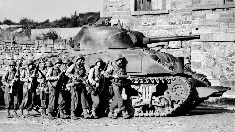 Taking shelter alongside an M4 Sherman medium tank, U.S. soldiers of the 60th Infantry Regiment advance into a Belgian town.