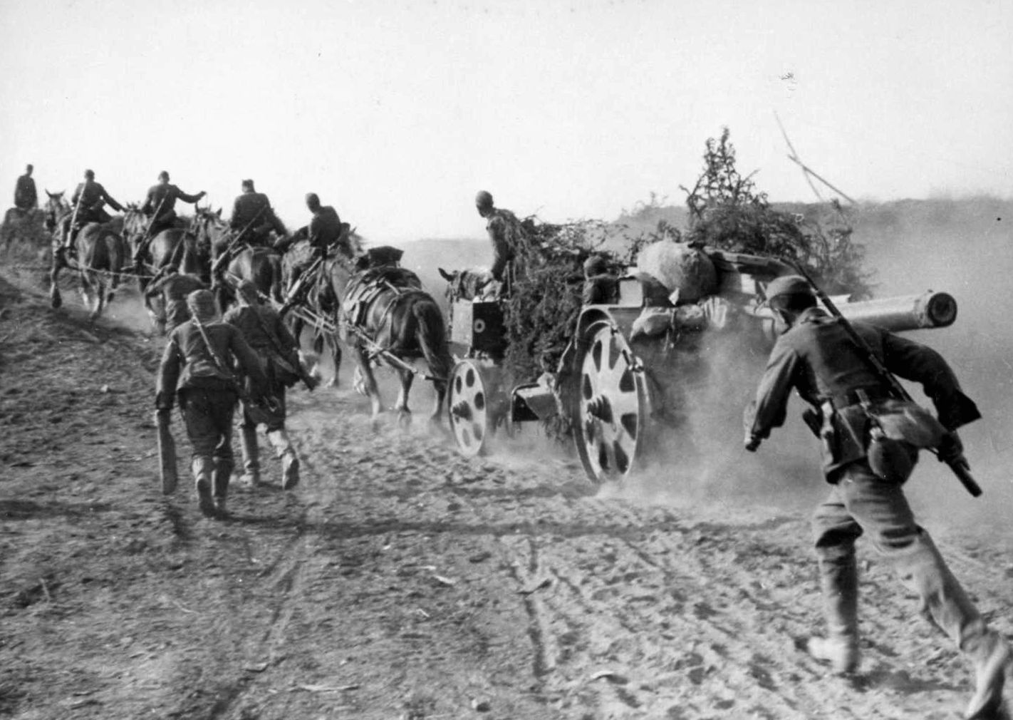 A team of horses strains under the weight of a heavy artillery piece, which produces a cloud of choking dust as German soldiers advance alongside. Note the makeshift attempt to camouflage the gun.
