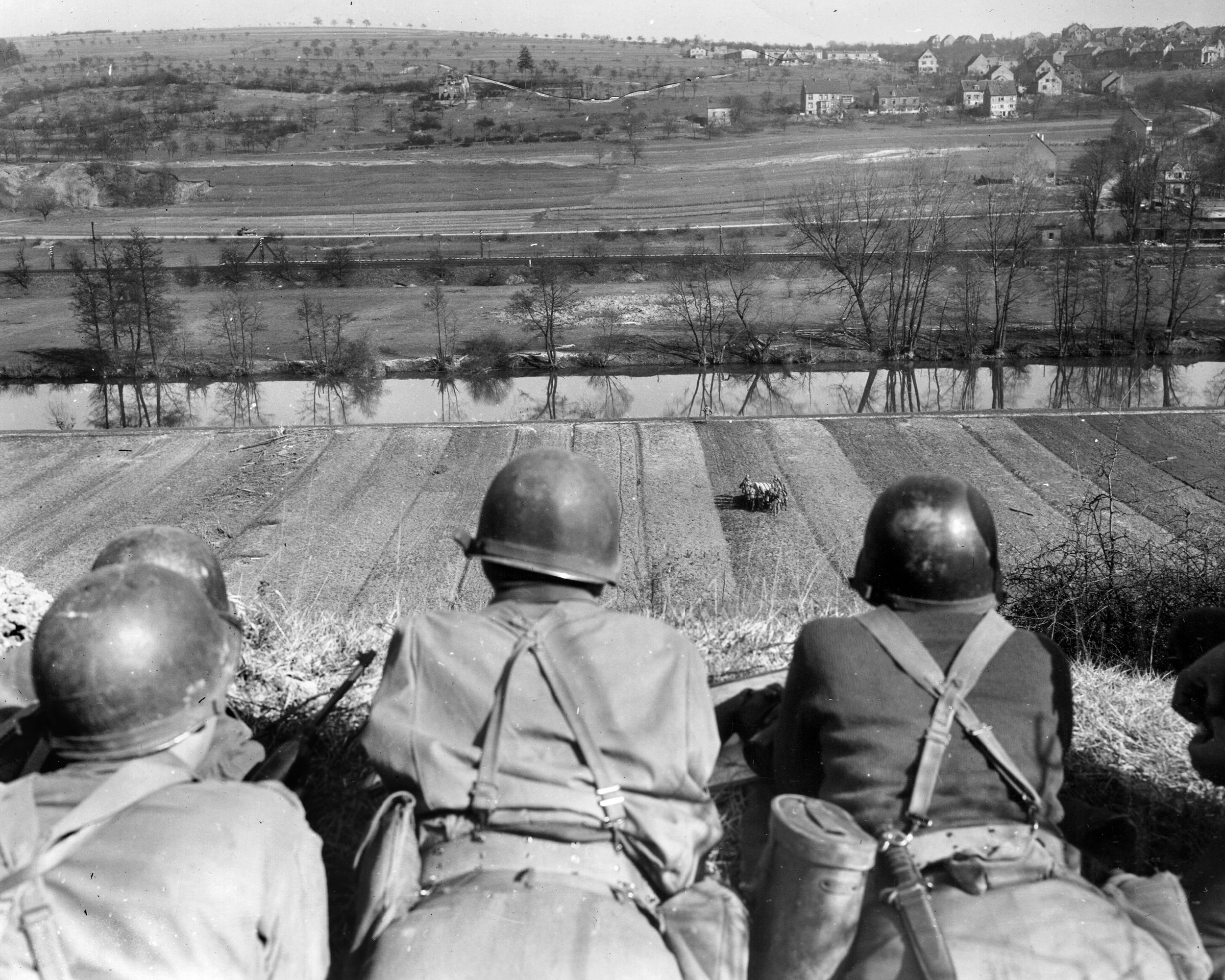 Observers on a bank overlooking the Saar River watch as men of the 3rd Battalion, 276th Regiment, carry a flat-bottom boat across a field toward the river, March 1945.