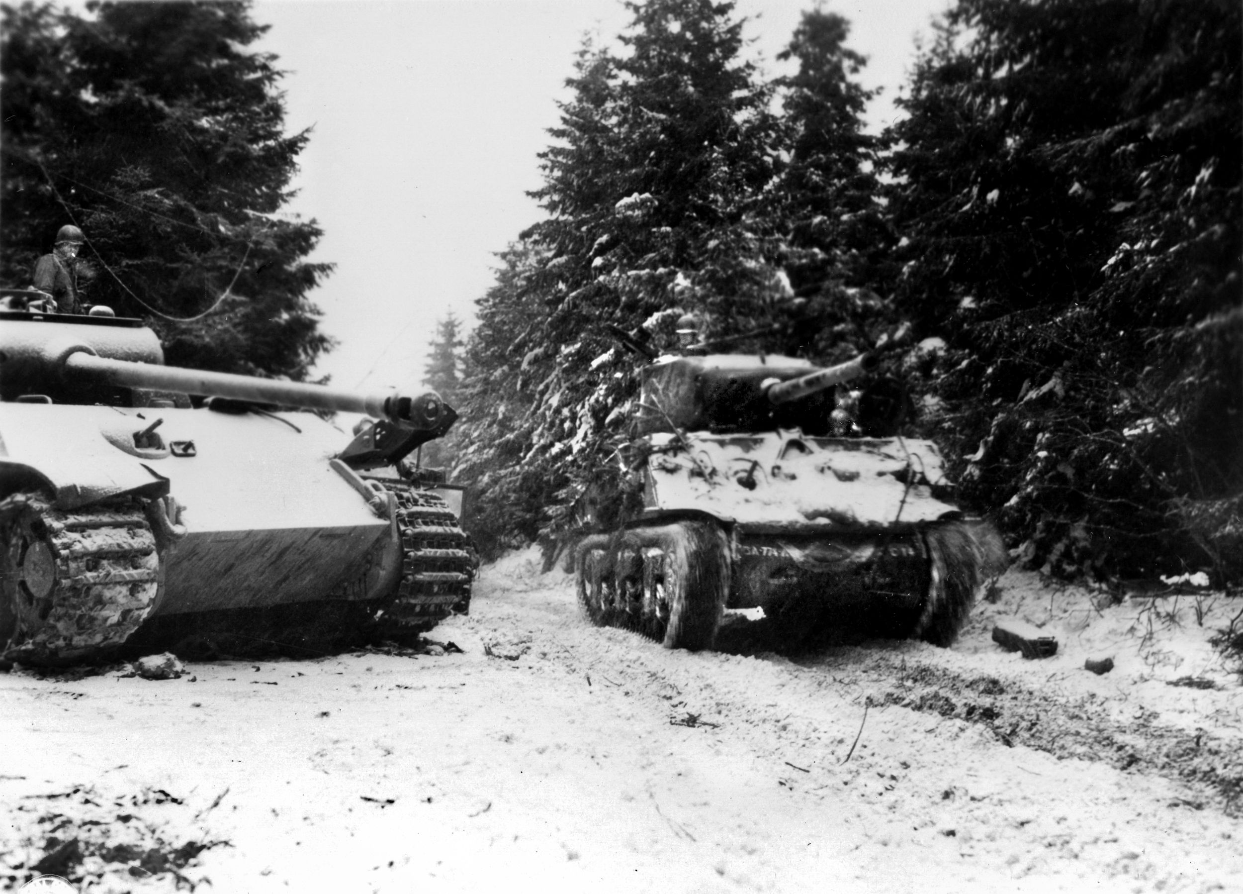A 3rd Armored Division Sherman (right) passes a knocked-out Pz.Kpfw V “Panther” tank on a snowy Belgian forest road during the Battle of the Bulge, December 1944. A curious GI can be seen examining the enemy tank.