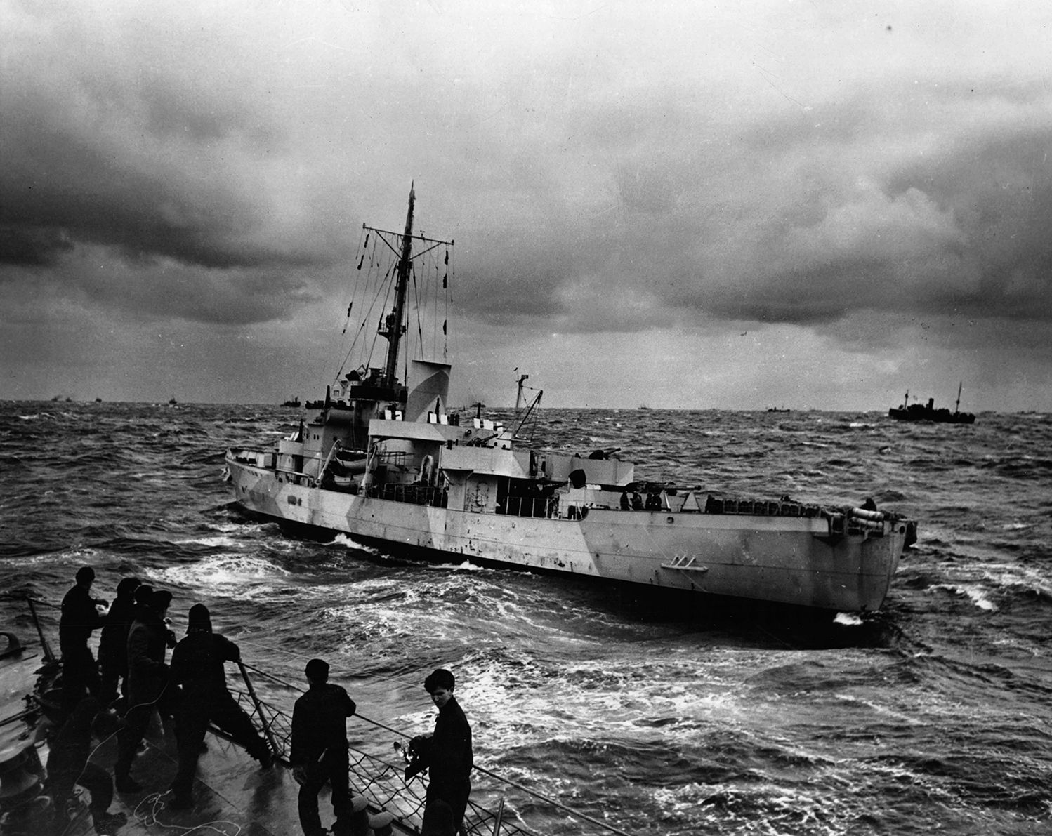 Its complement of sailors at general quarters and its decks laden with depth charges, a U.S. Coast Guard cutter rides swells in the Atlantic Ocean while on convoy escort duty, protecting merchant ships loaded with supplies and equipment for the war in Europe.
