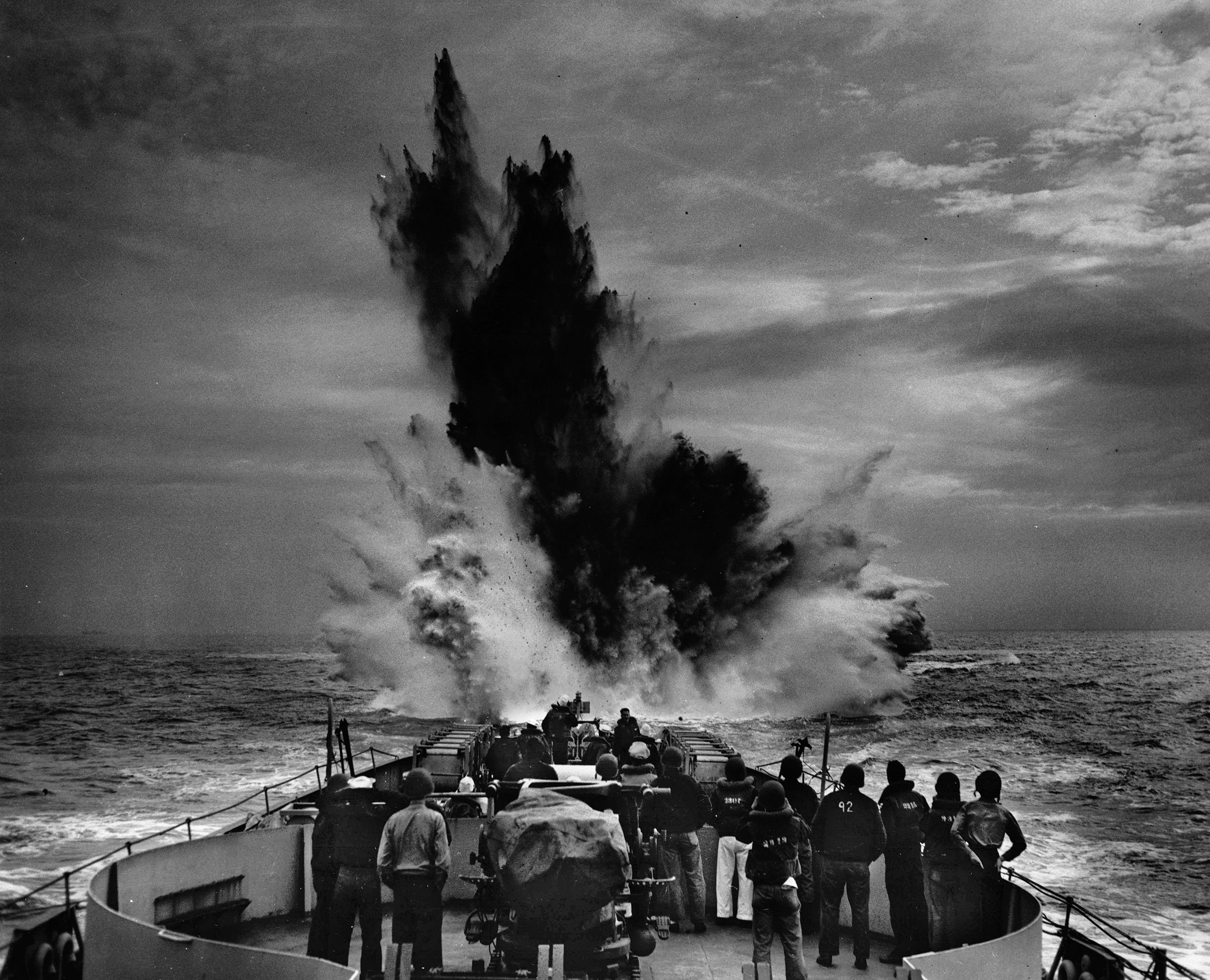 A depth charge explodes beyond the stern of a Coast Guard cutter in the Atlantic while crewmen watch the resulting cascade of seawater. Coast Guard cutters played a key role in the fight against German U-boats.