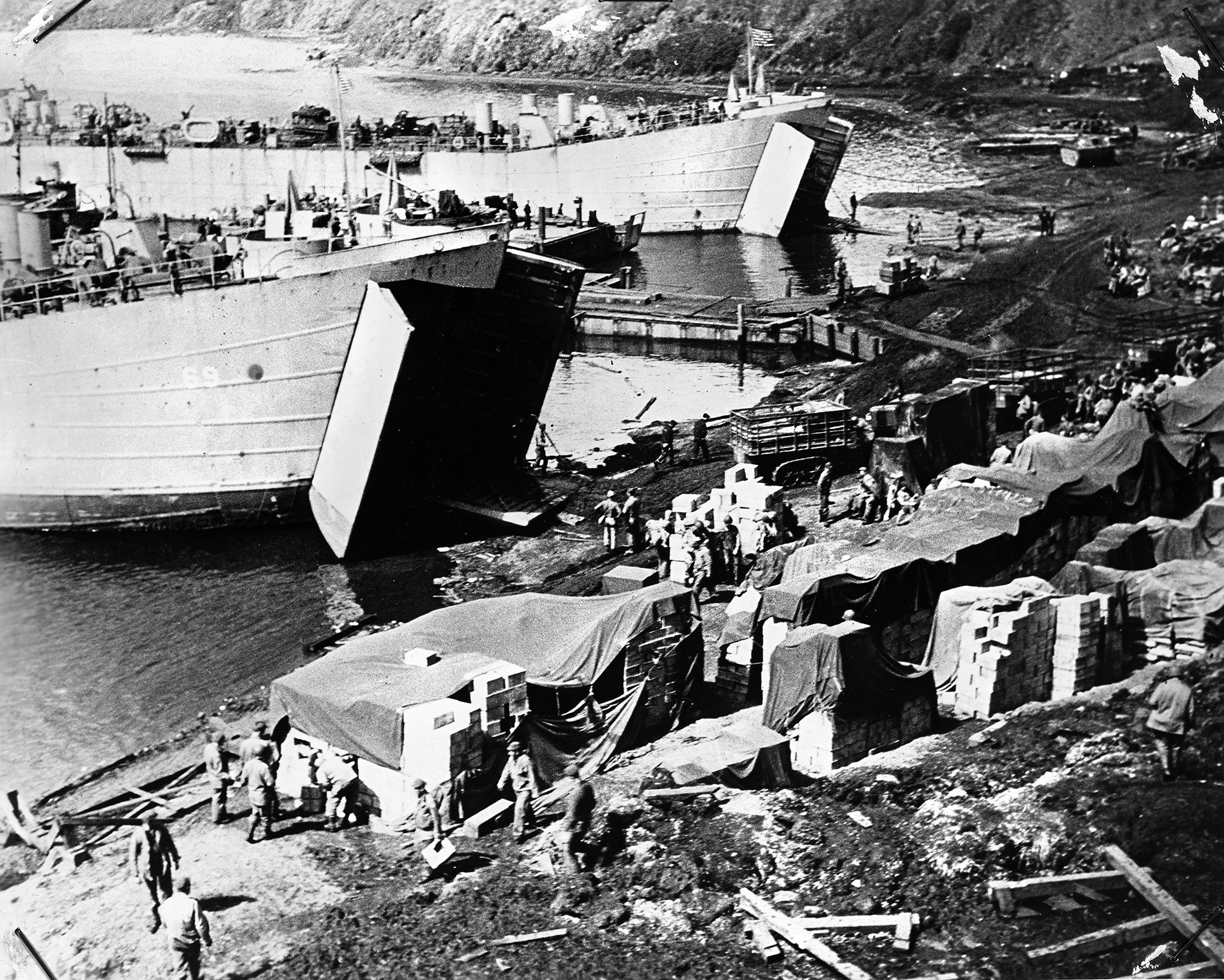 Coast Guardsmen and soldiers of the U.S. Army unload equipment and supplies on a beach in support of Allied landings. Note the large swinging doors of the LSTs (Landing Ship, Tank), which allowed the vessels to transport heavy loads, including tanks and other armored vehicles.