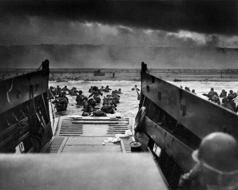 A Coast Guard crewman aboard a landing craft is shown at lower right as his human cargo of combat troops exits toward the Easy Red sector of Omaha Beach in Normandy on D-Day. This photo was taken while the Americans were under tremendous German fire from the bluffs visible in the distance.