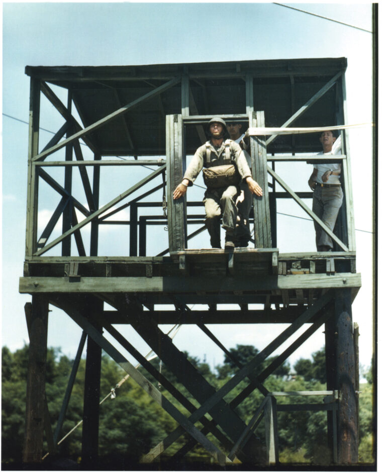 A paratrooper prepares to jump from a mock tower while instructors look on. Students were taught to put their hands outside the plane door before jumping. If they kept their hands inside, the instructors would push them aside and let the other students jump.