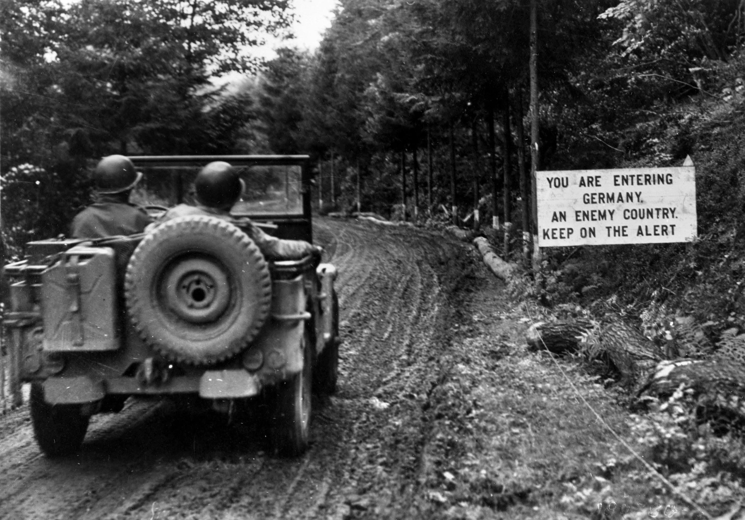 An American Jeep rolls down a muddy road into Germany, passing a sign that is both a greeting and a warning. The U.S. First Army, under the command of General Courtney Hodges, entered Germany initially in November 1944, during the fighting in the Hurtgen Forest.